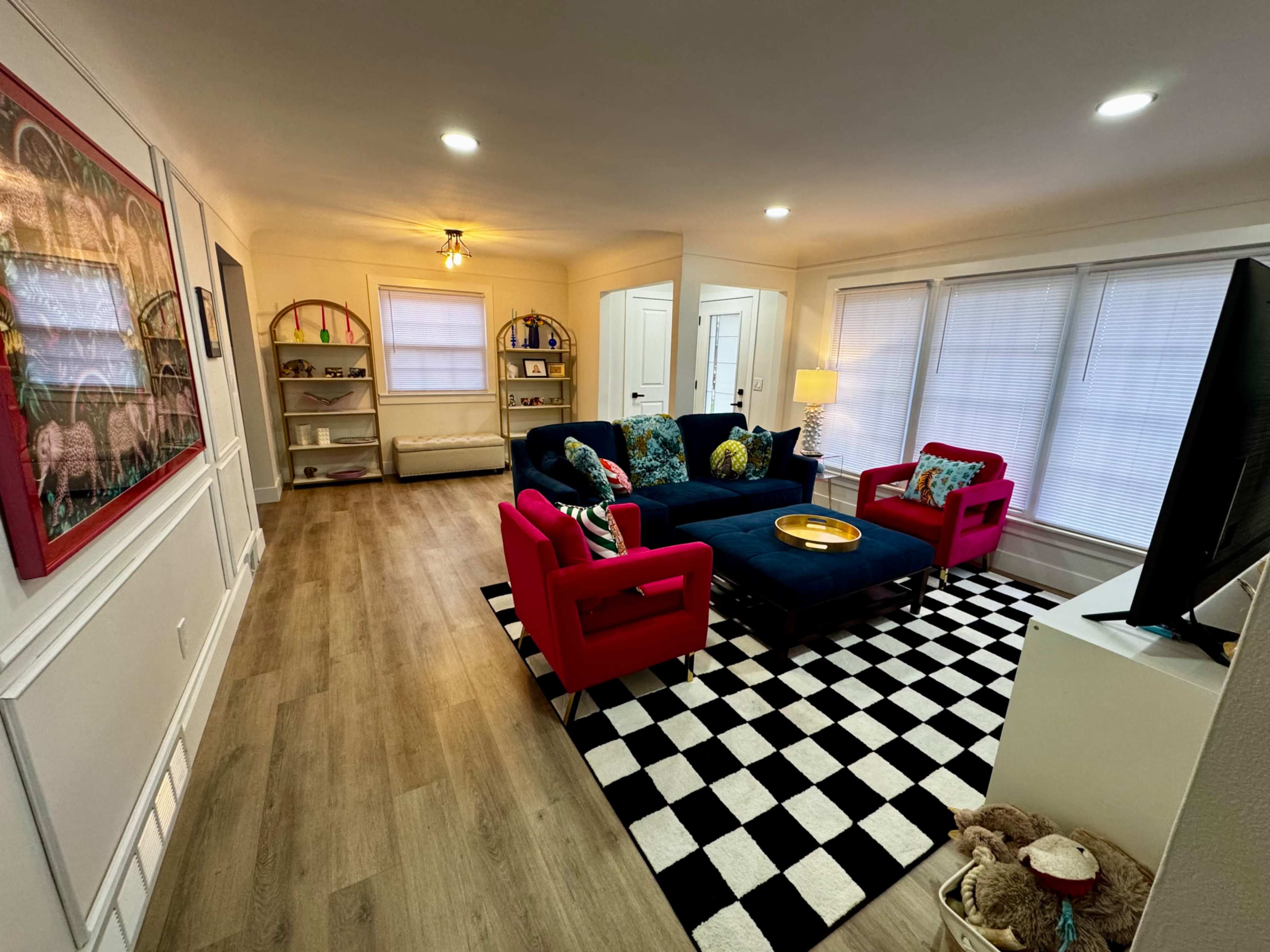 The image shows a brightly colored living room with a navy blue sofa, red chairs, a checkered black-and-white rug, and illuminated by natural light through large windows.