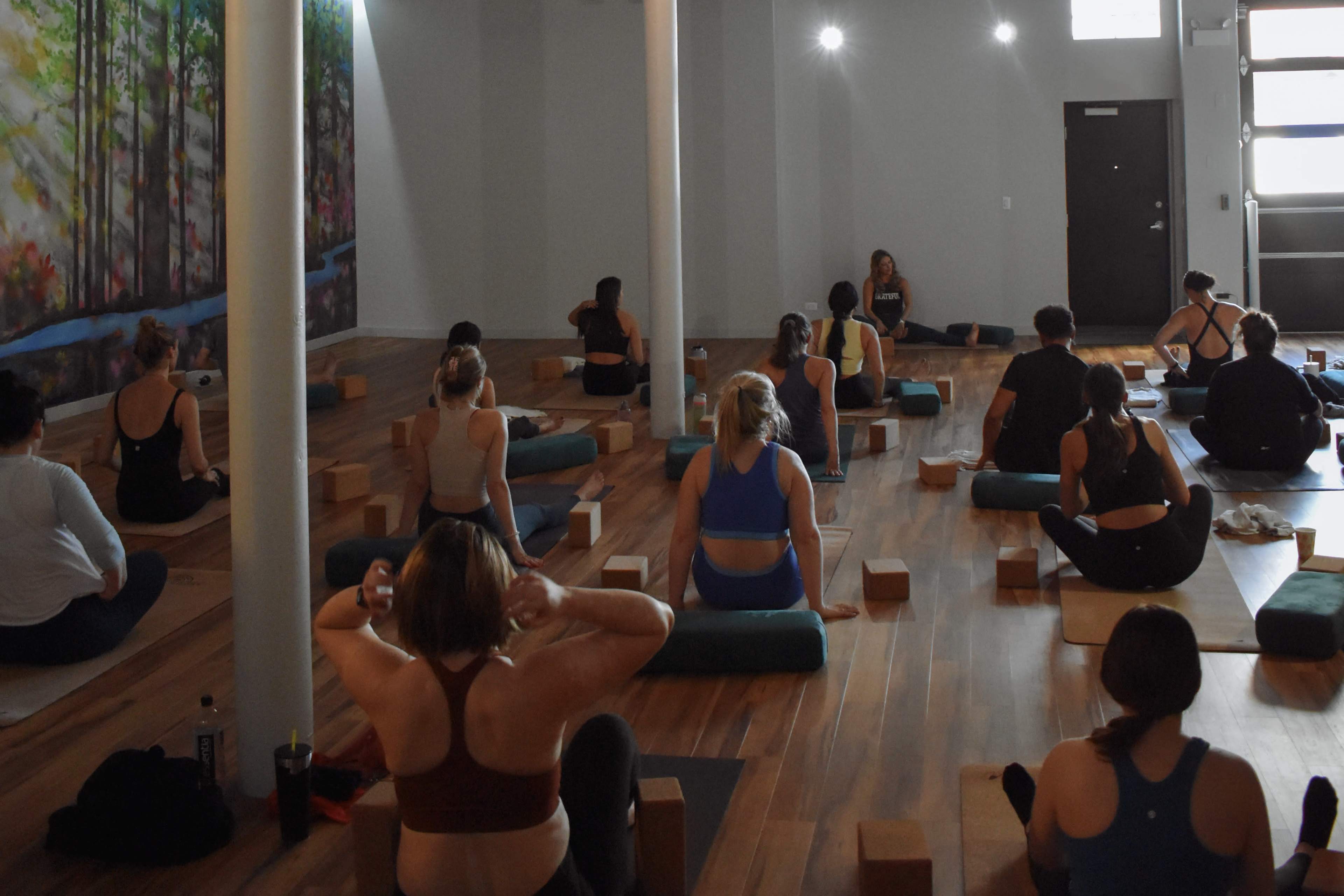 A group of individuals participates in a yoga class in a studio with wooden floors and a mural depicting trees.