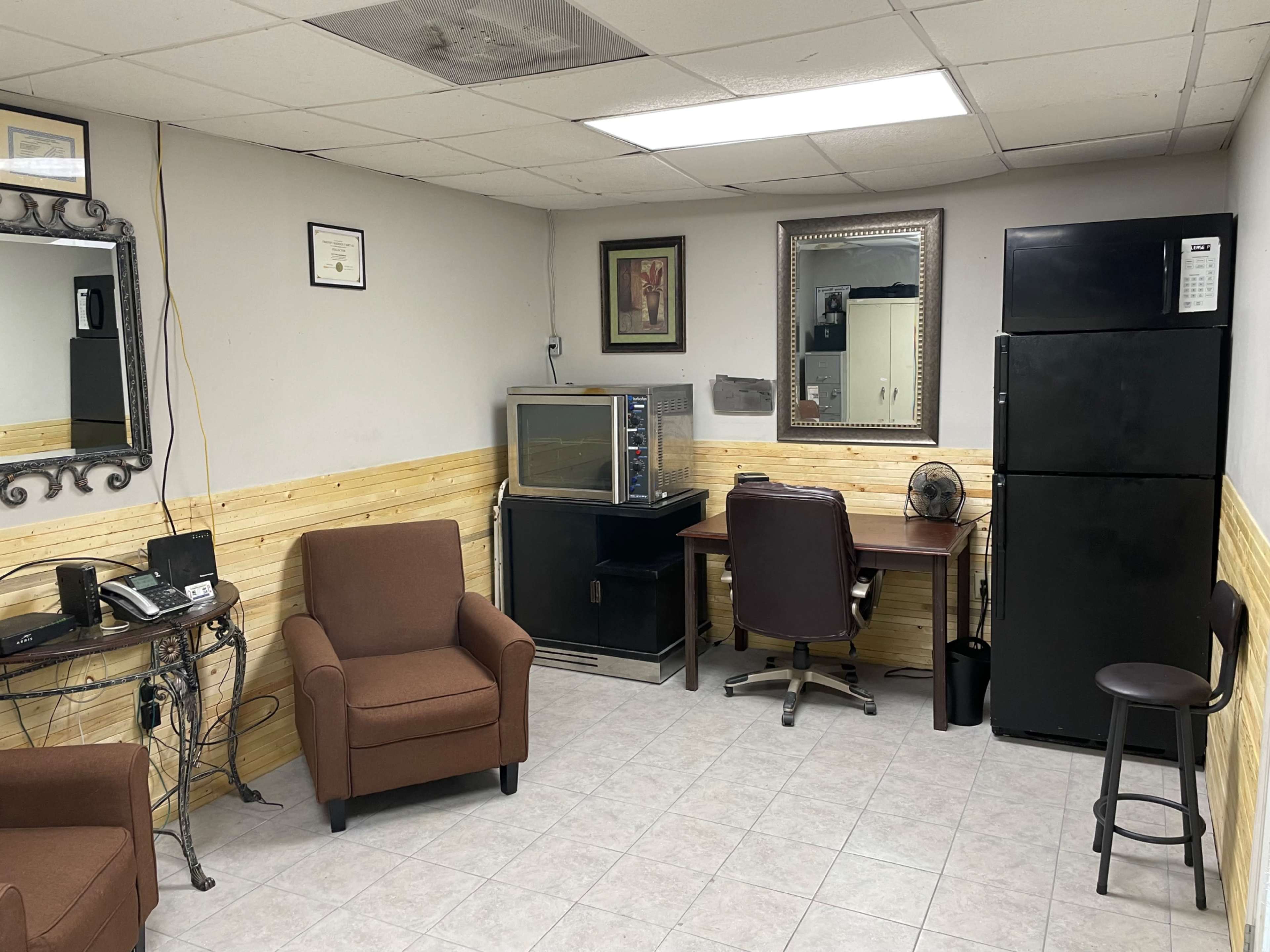 A small office space with two brown armchairs, a desk with a fan, a microwave, a black refrigerator, and a wall-mounted mirror.