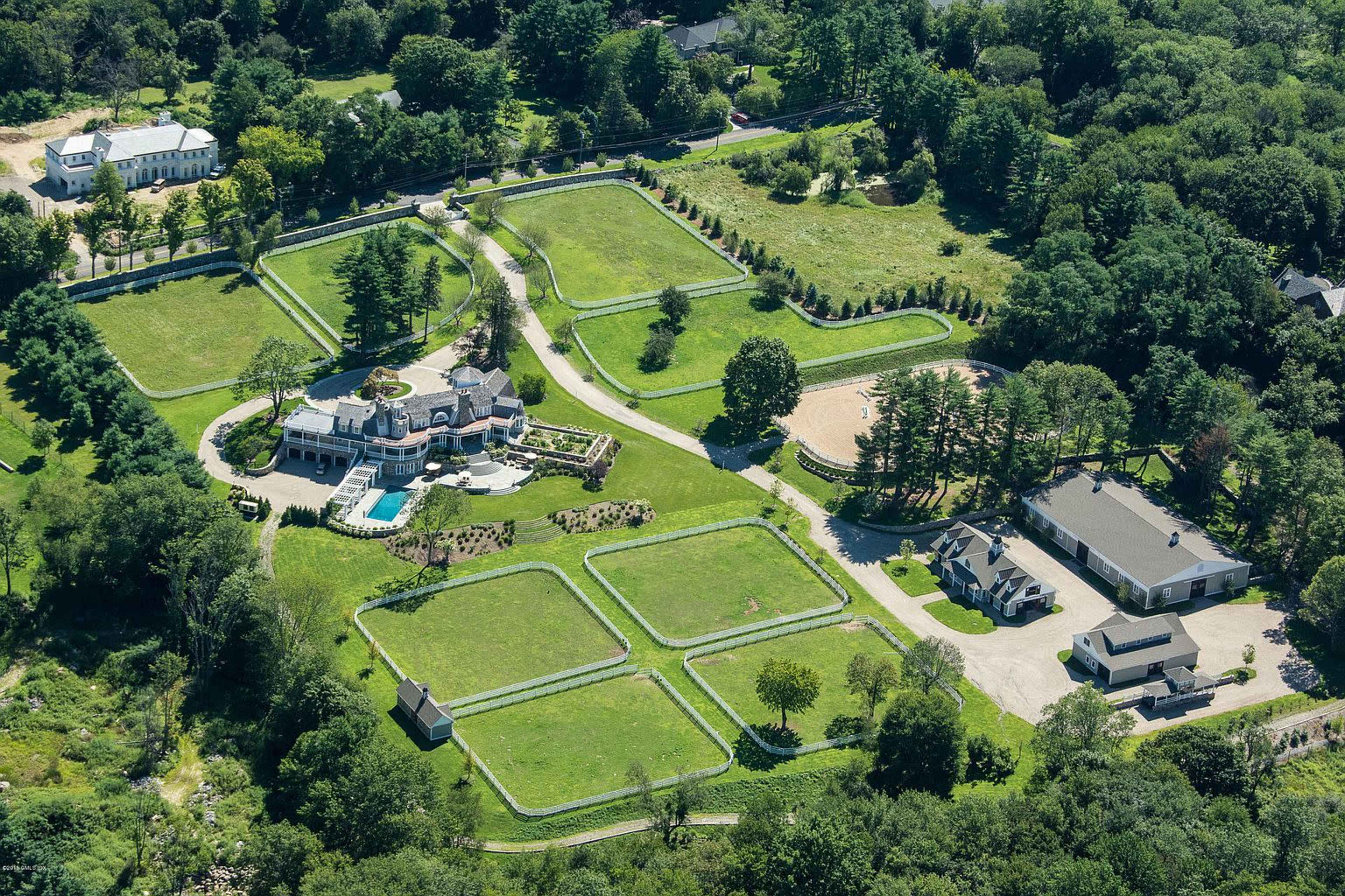 The image shows a large estate with manicured lawns, a swimming pool, and several buildings situated on expansive green grounds bordered by trees.