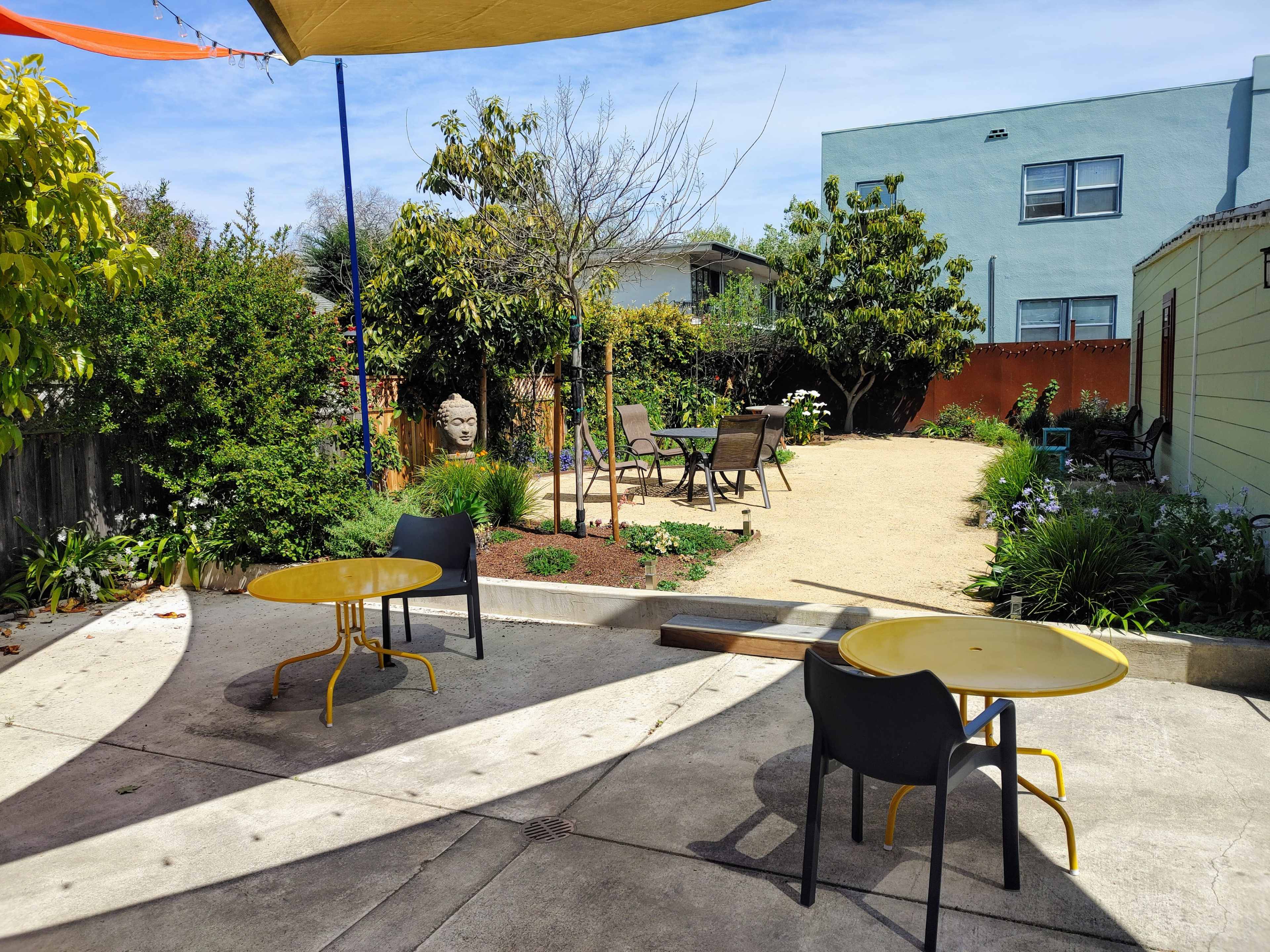 A courtyard features two yellow tables and chairs with a gravel path leading to a landscaped garden filled with greenery and seating areas.
