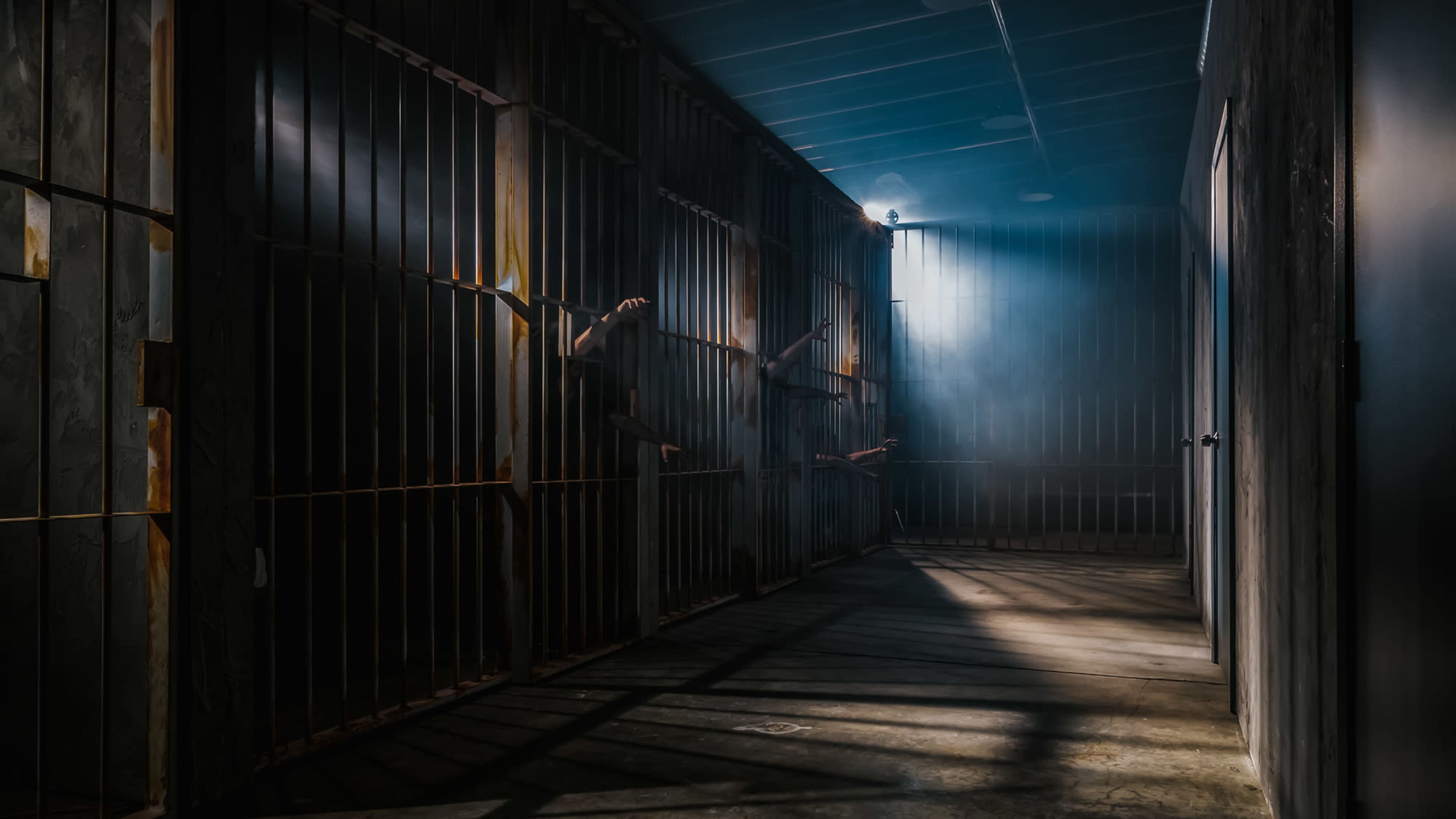 A dimly lit hallway features several jail cells with shadows of hands reaching out through the bars.