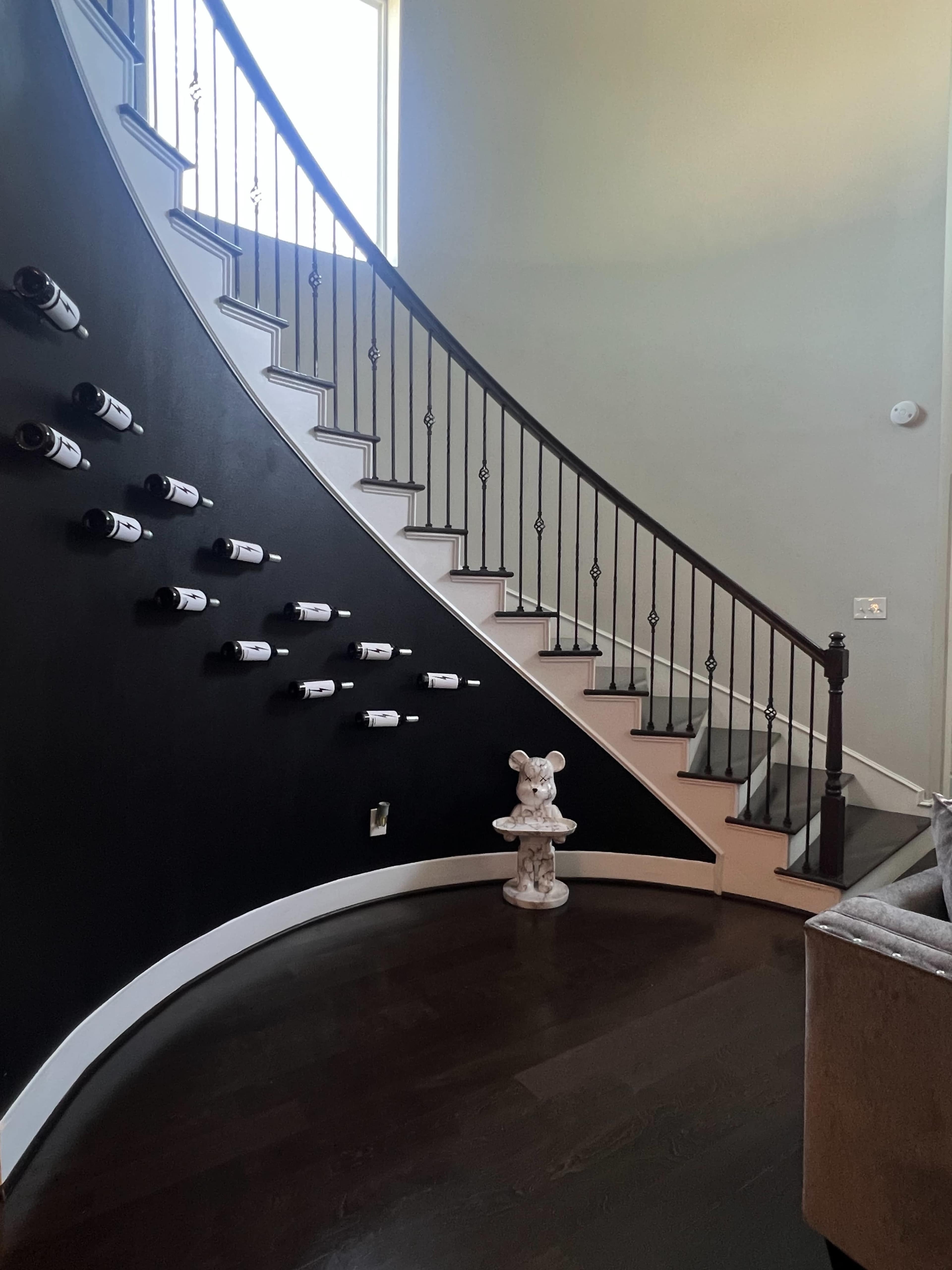 A curved staircase with a black wall displays wine bottles, while a decorative statue stands at the bottom.
