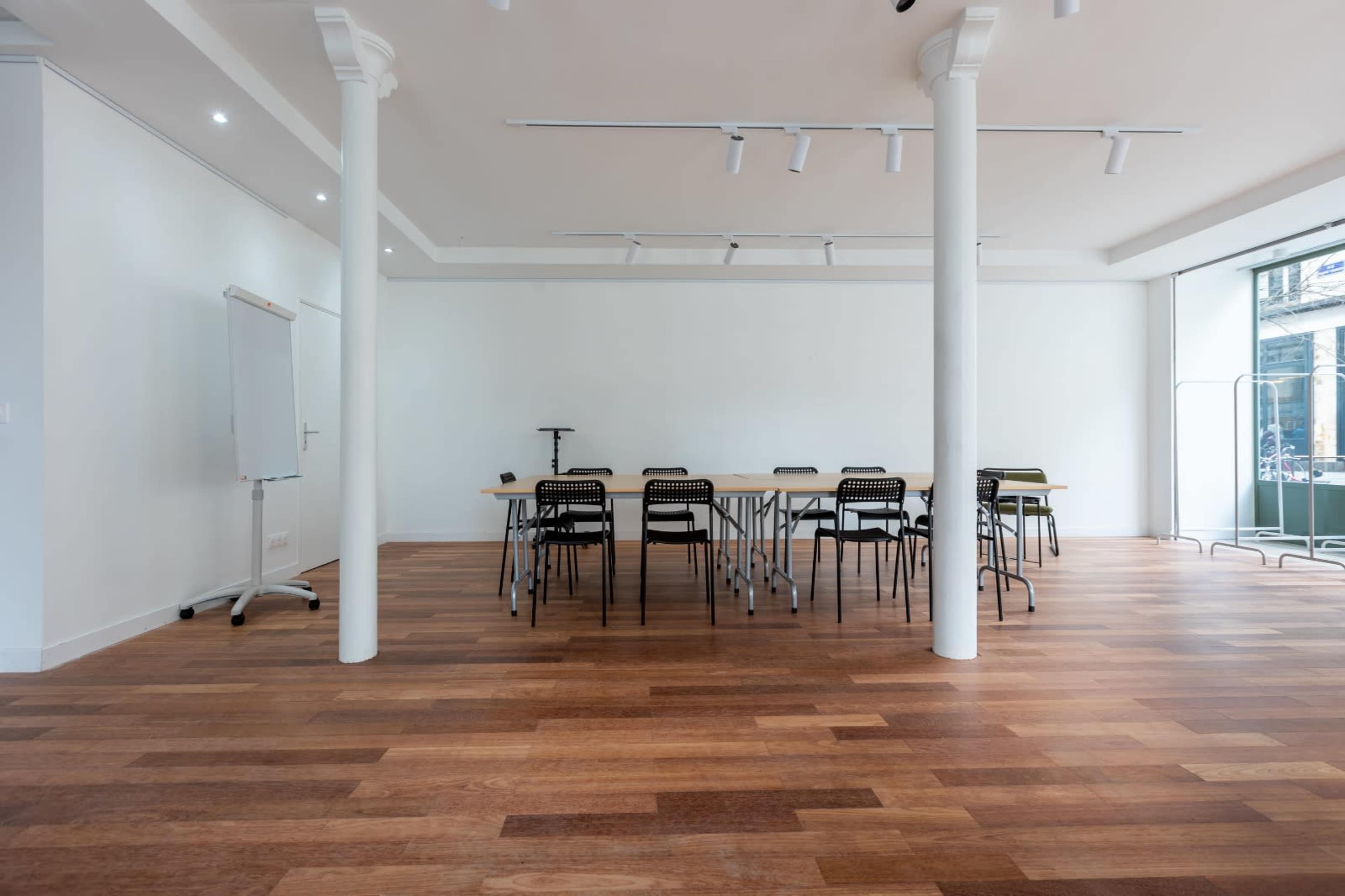 A spacious, well-lit meeting room features a long table with black chairs arranged around it, and large windows allowing natural light to enter.