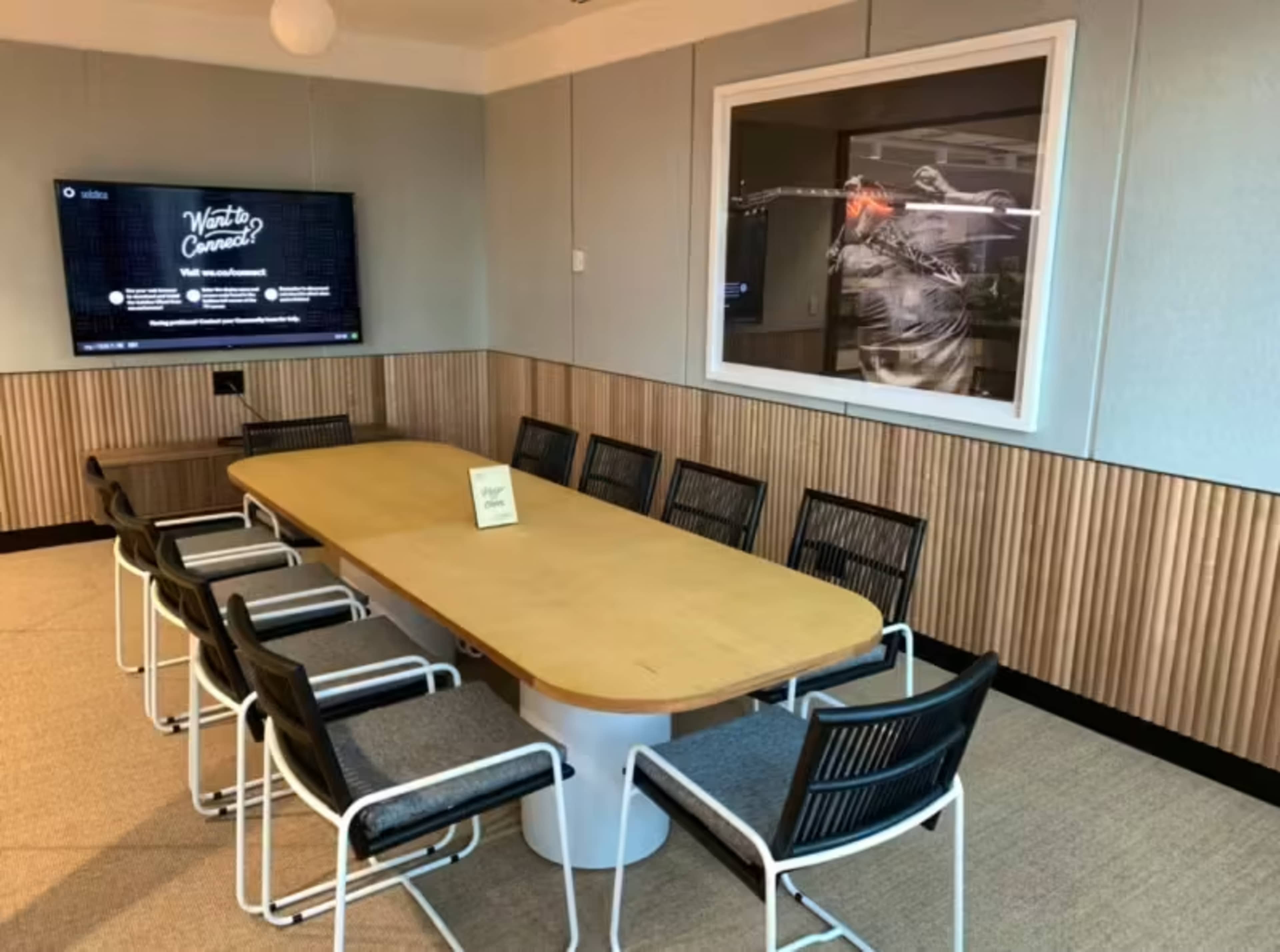 A large conference room features a long wooden table surrounded by several chairs, with a television mounted on the wall and a framed picture hanging nearby.