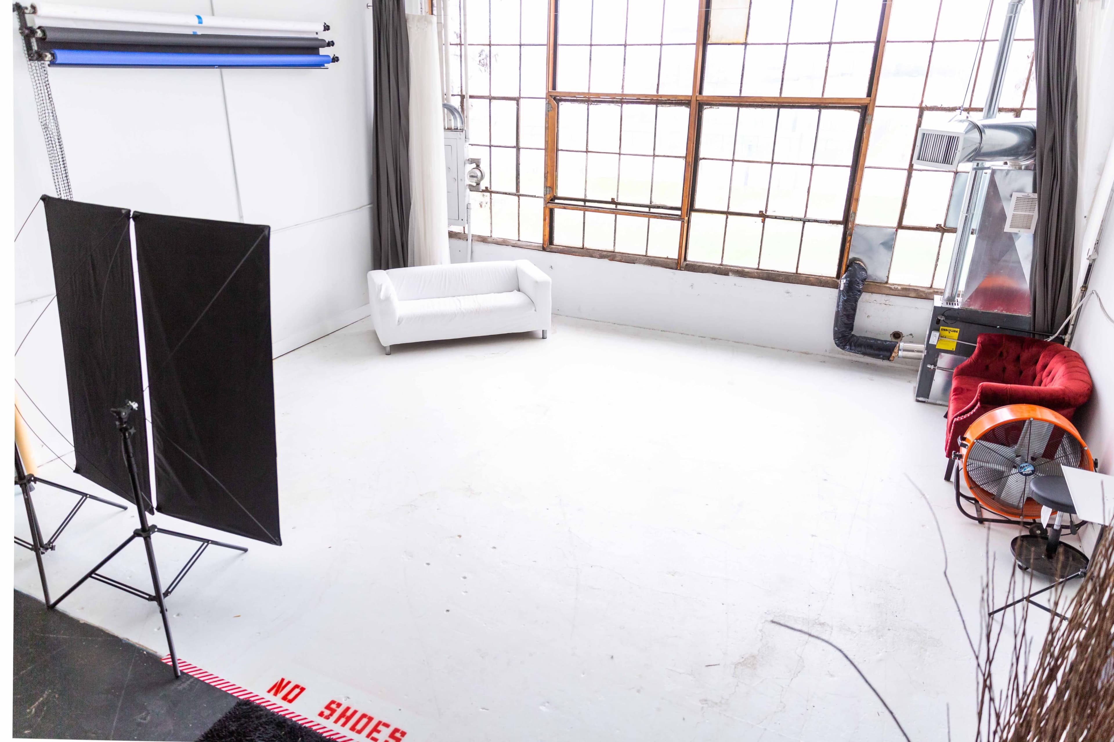 The image shows a spacious, well-lit studio with large windows, a white couch, a red chair, and photography backdrops.