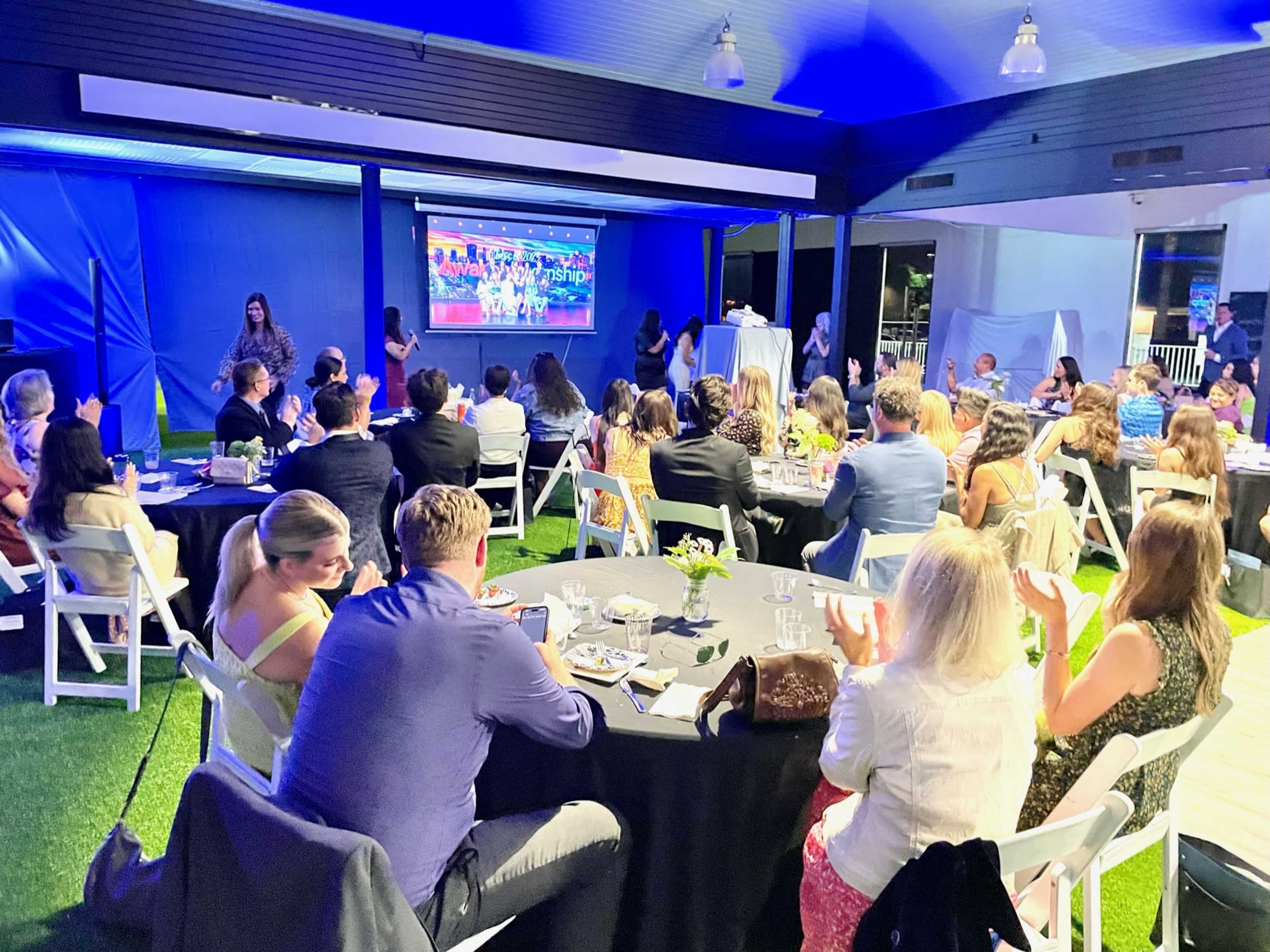 A large group of people is seated at round tables, watching a presentation on a screen against a backdrop of blue lighting.