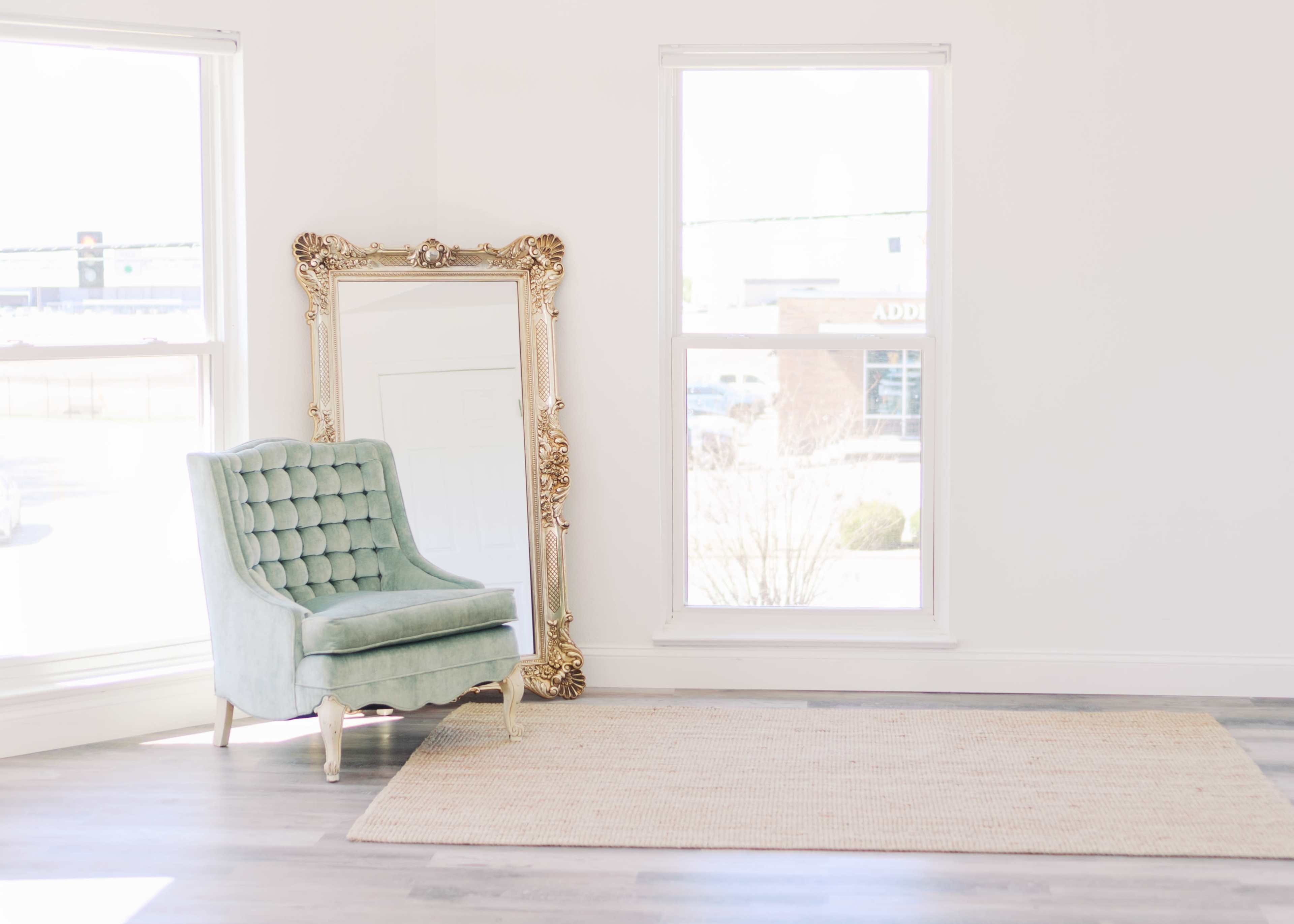 The image shows a light-colored room featuring a blue-green upholstered chair next to a large ornate mirror, with two windows allowing natural light to enter.