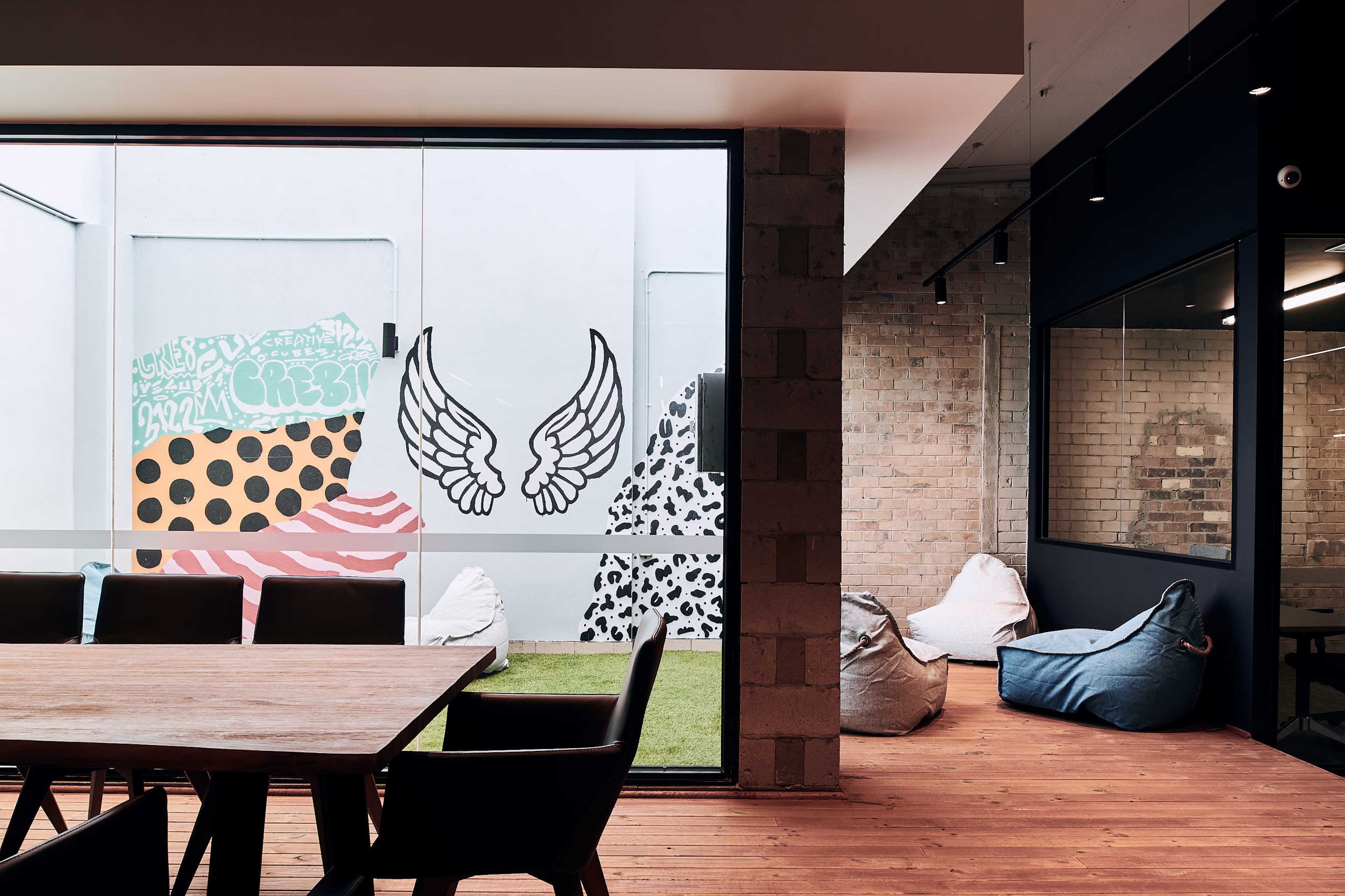 The image shows a modern office space featuring a glass wall with colorful wall art, including abstract shapes and wings, alongside a wooden table and bean bag seating in a common area.