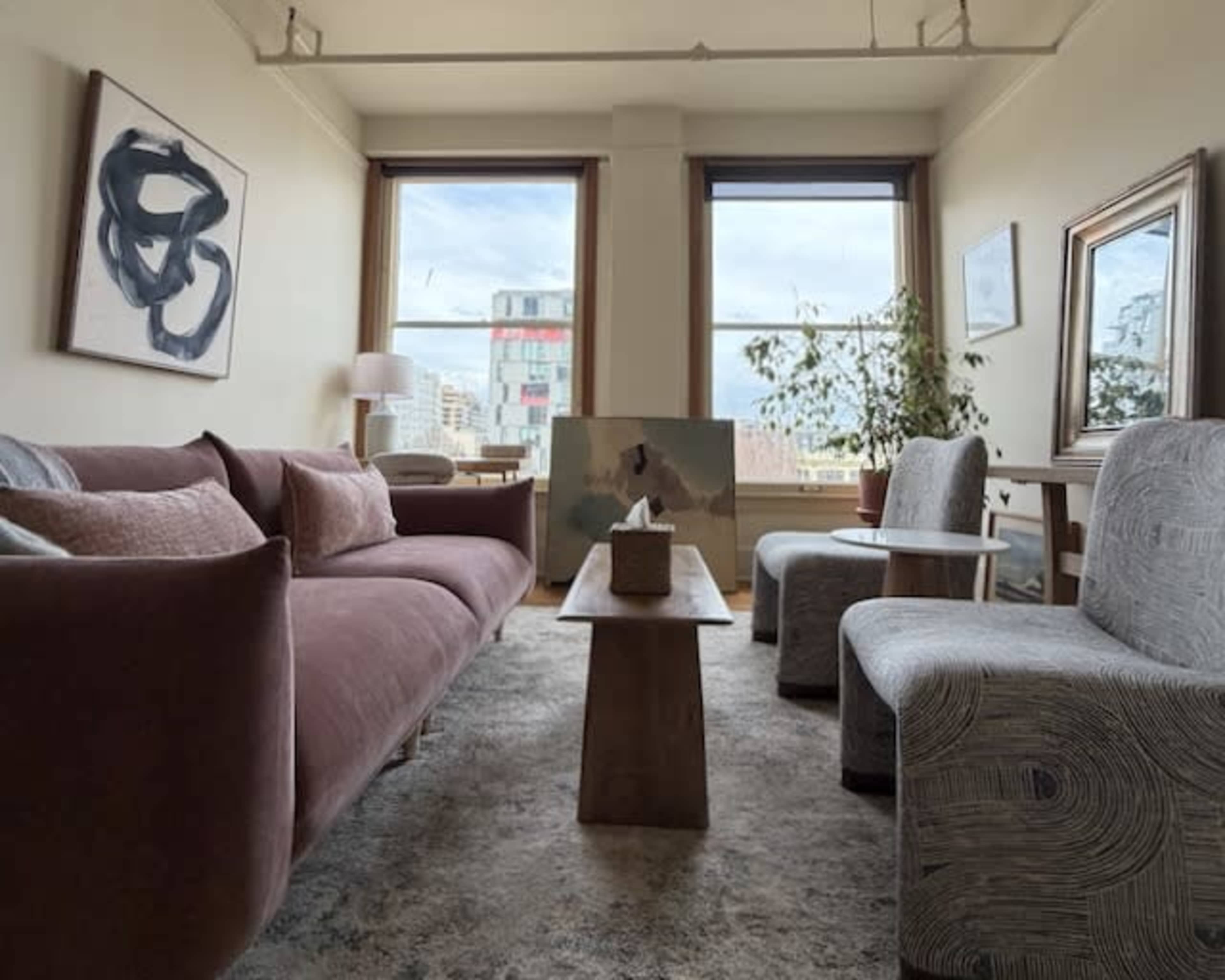 The image shows a modern living room with a pink sofa, two patterned armchairs, a wooden coffee table, and large windows revealing an urban view.