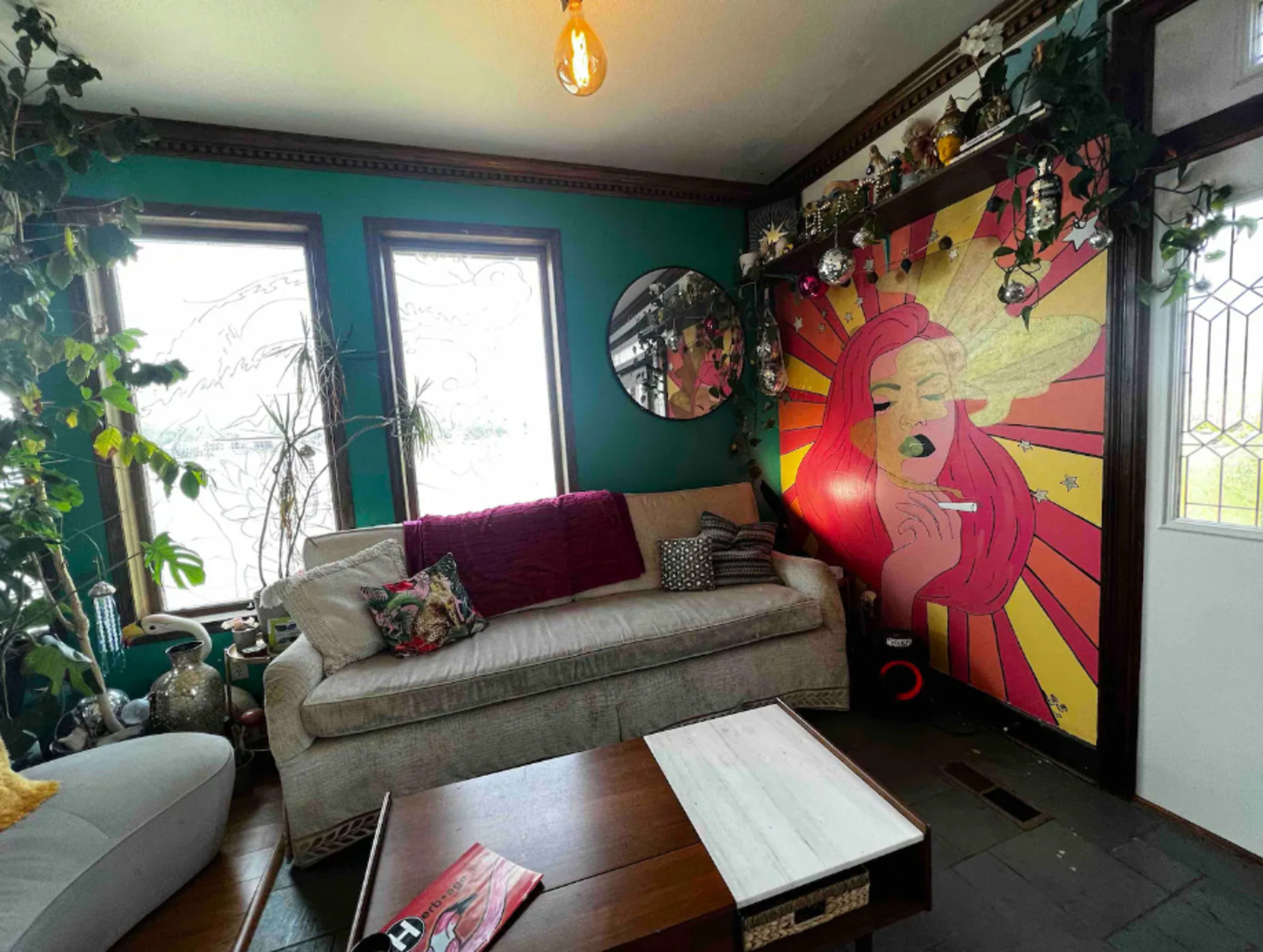 The image shows a living room with a beige sofa, a wooden coffee table, and a vibrant mural of a woman against a colorful backdrop on one wall, surrounded by plants and large windows.