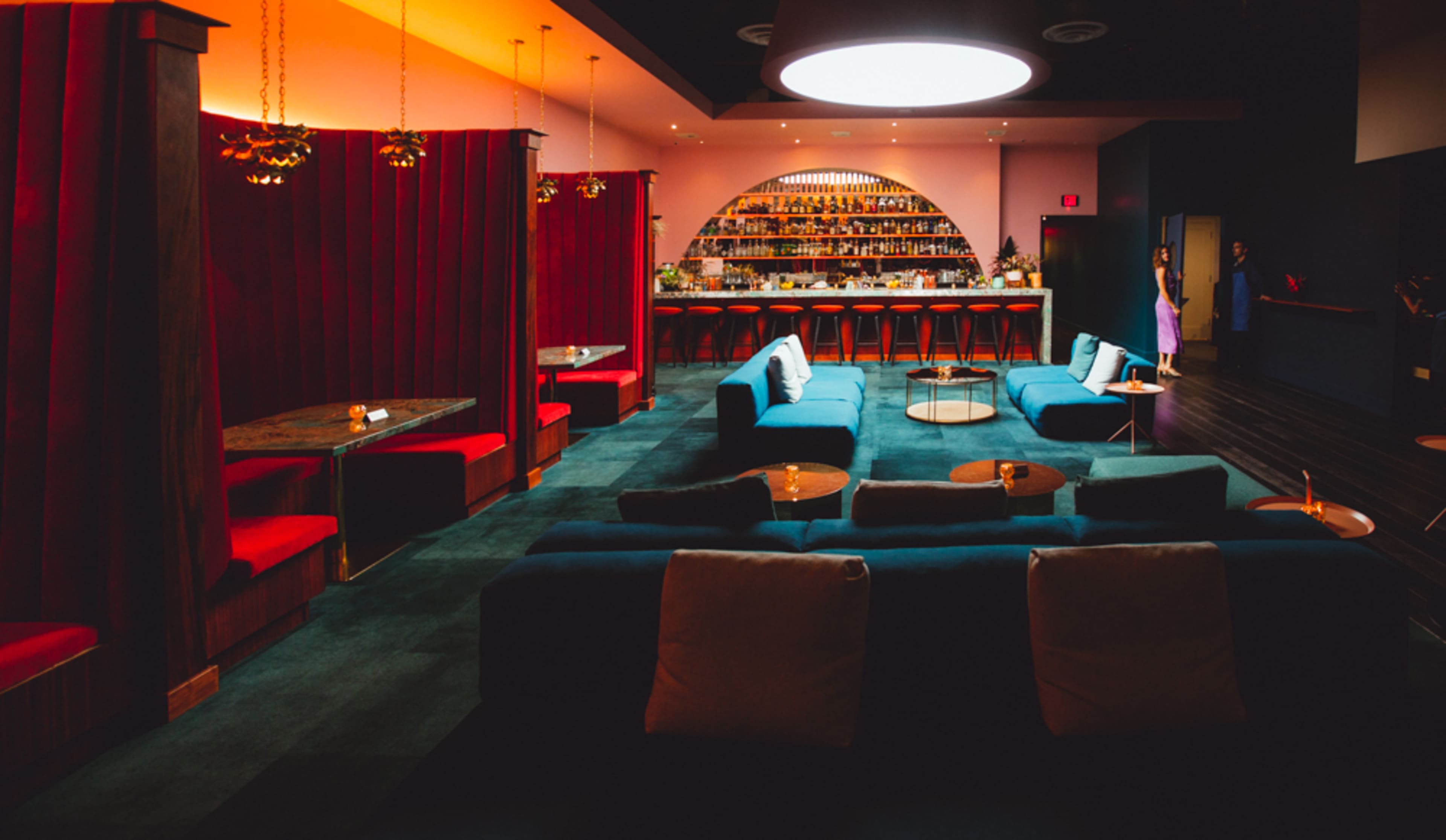 A stylish bar lounge featuring red and blue seating, a well-stocked bar in the background, and warm lighting.