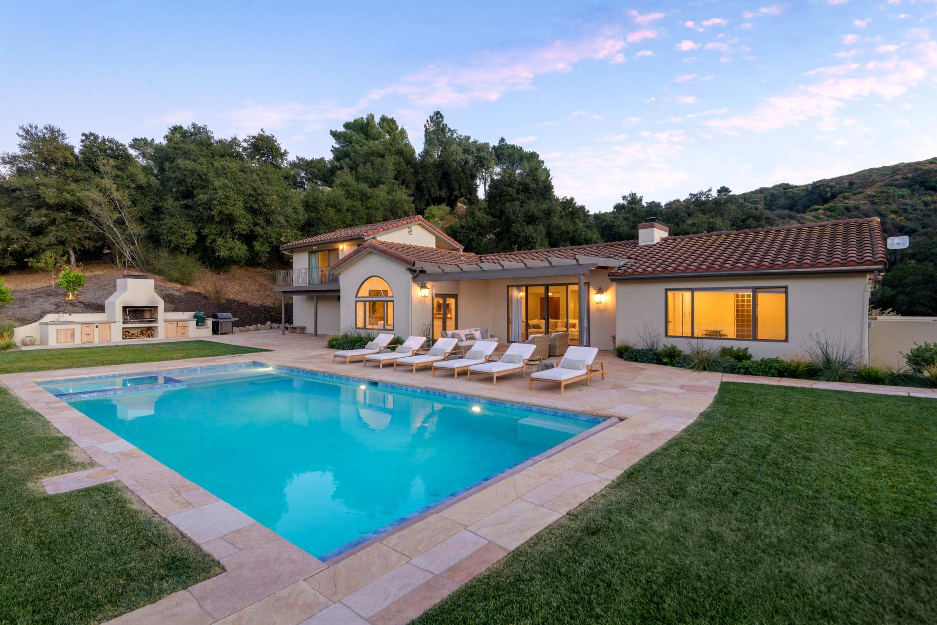 A modern two-story house with a swimming pool and lounge chairs is situated in a landscaped backyard surrounded by trees.