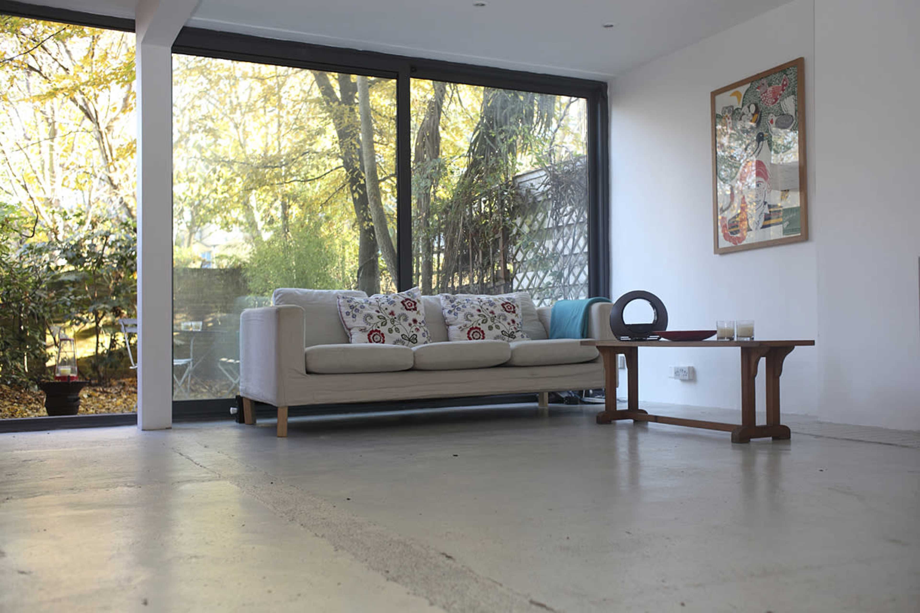 A modern living room features a light-colored sofa with patterned cushions and a wooden side table, alongside large windows overlooking a garden.
