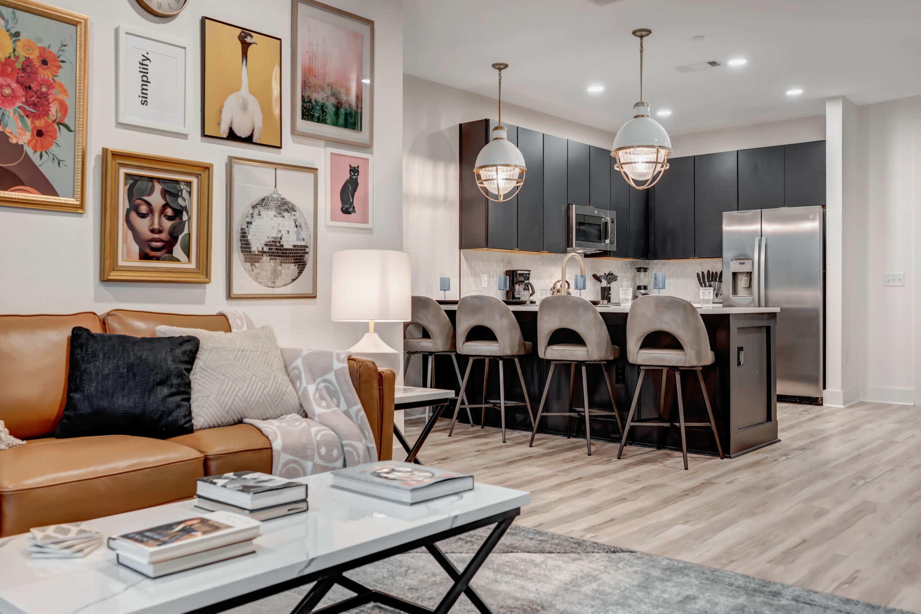 A modern living room and kitchen feature an open layout with a brown leather sofa, a coffee table, and a bar seating area with gray stools.