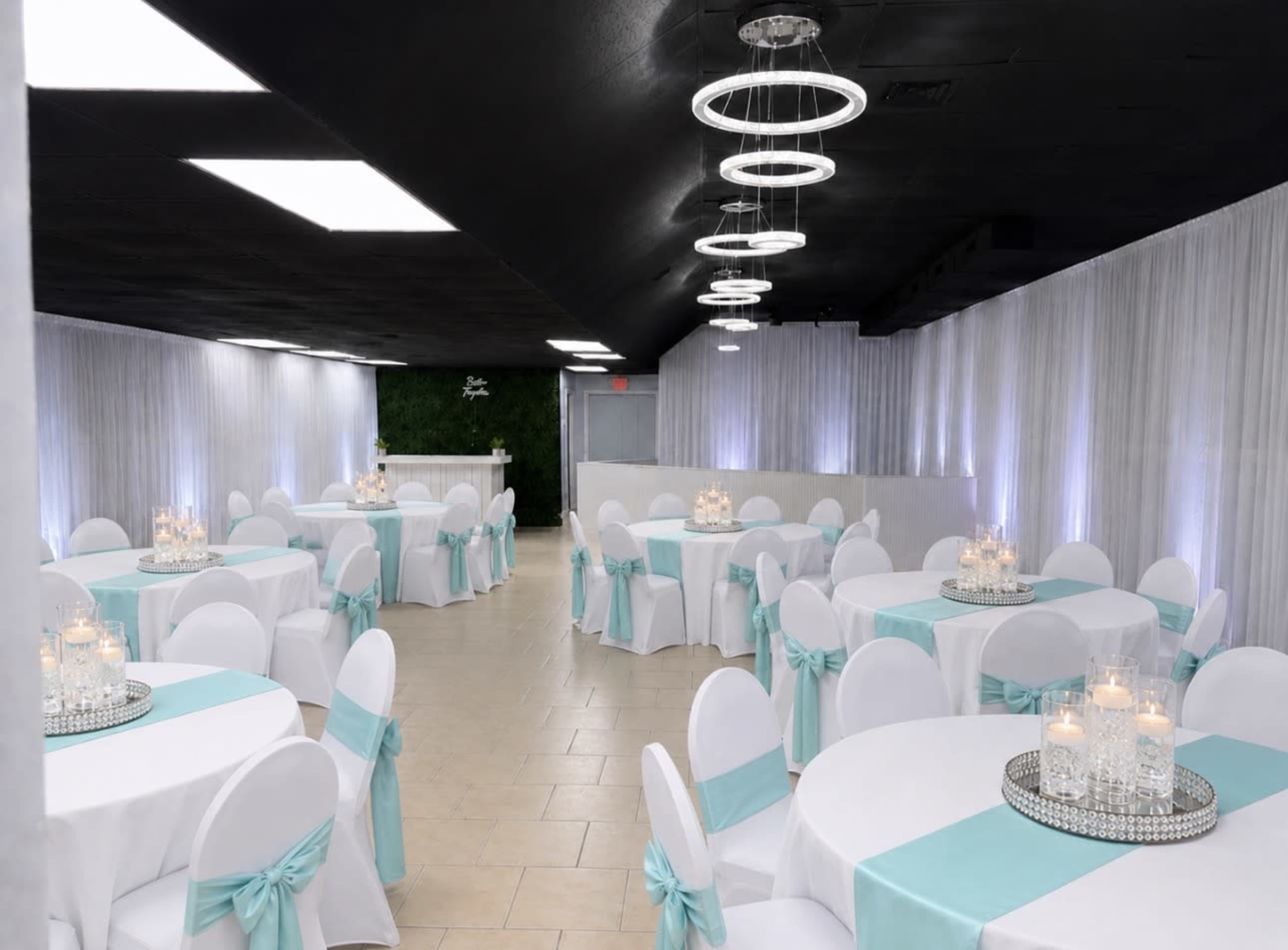 The image shows a banquet hall set up with circular tables covered in white tablecloths and adorned with light blue sashes on the chairs, illuminated by soft lighting.