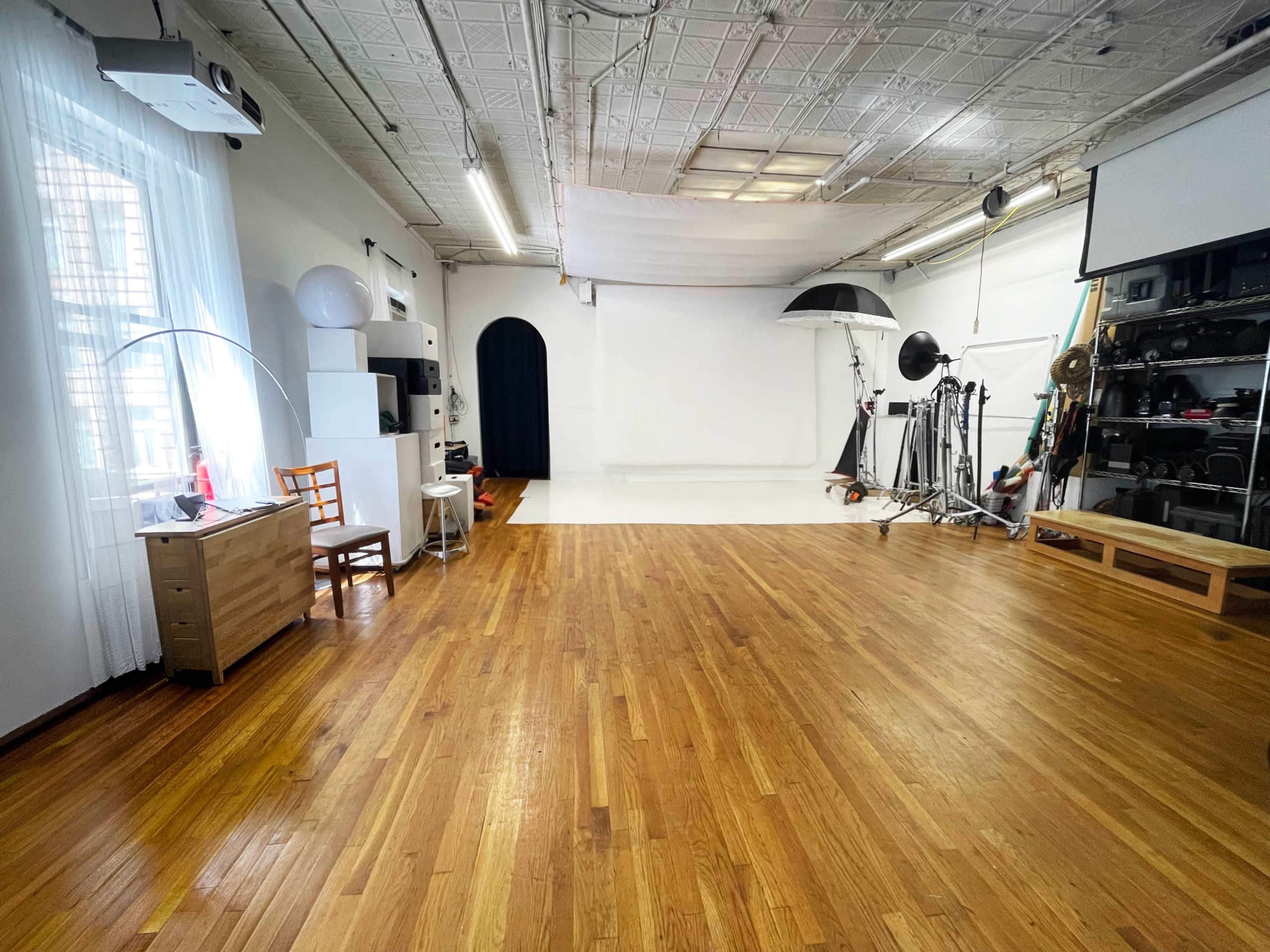 The image depicts a spacious photography studio with wooden floors, white walls, and various lighting equipment positioned around a central empty backdrop.