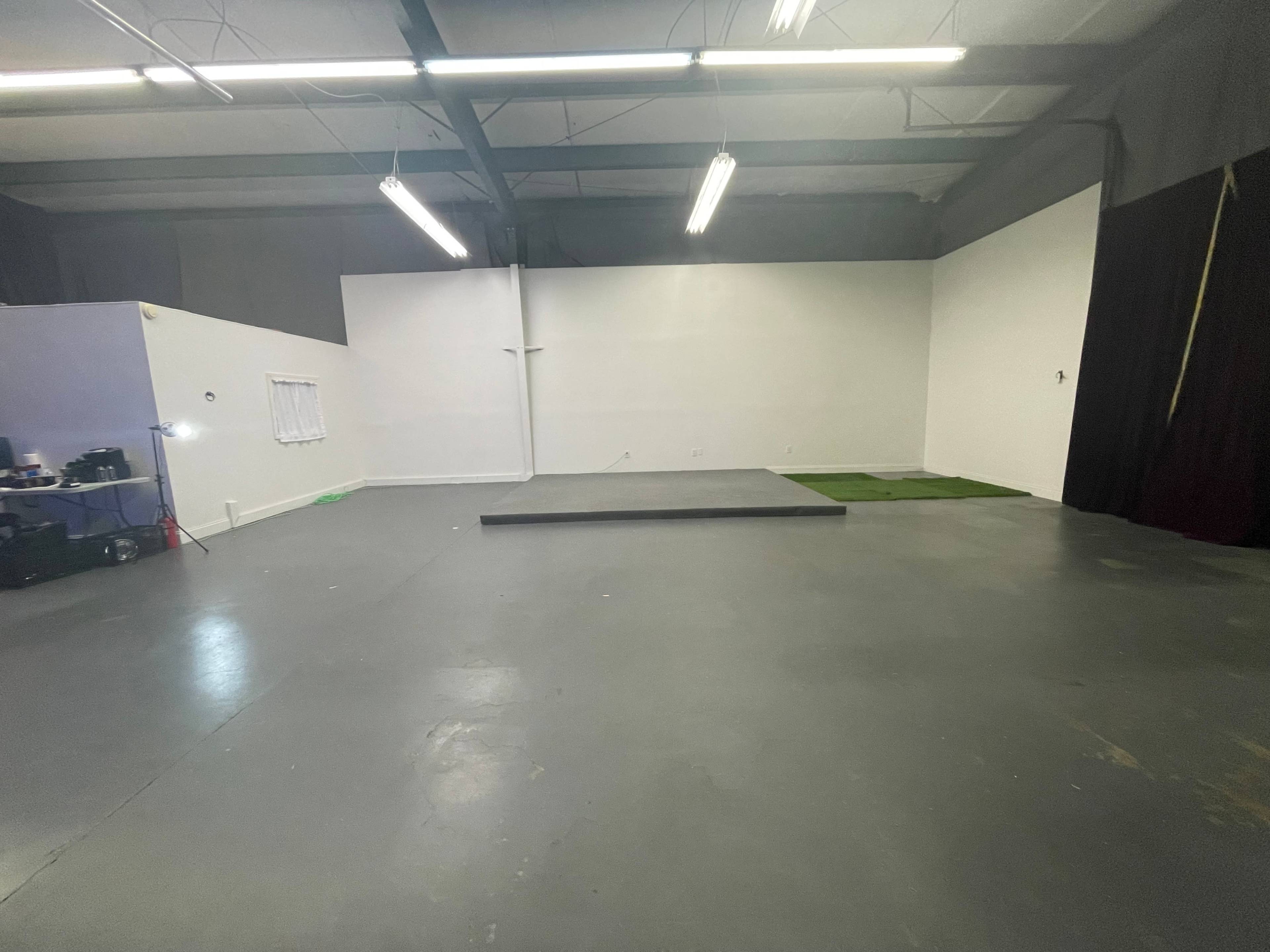 An empty room with a gray floor, a raised platform at the center, and white walls, illuminated by overhead fluorescent lights.