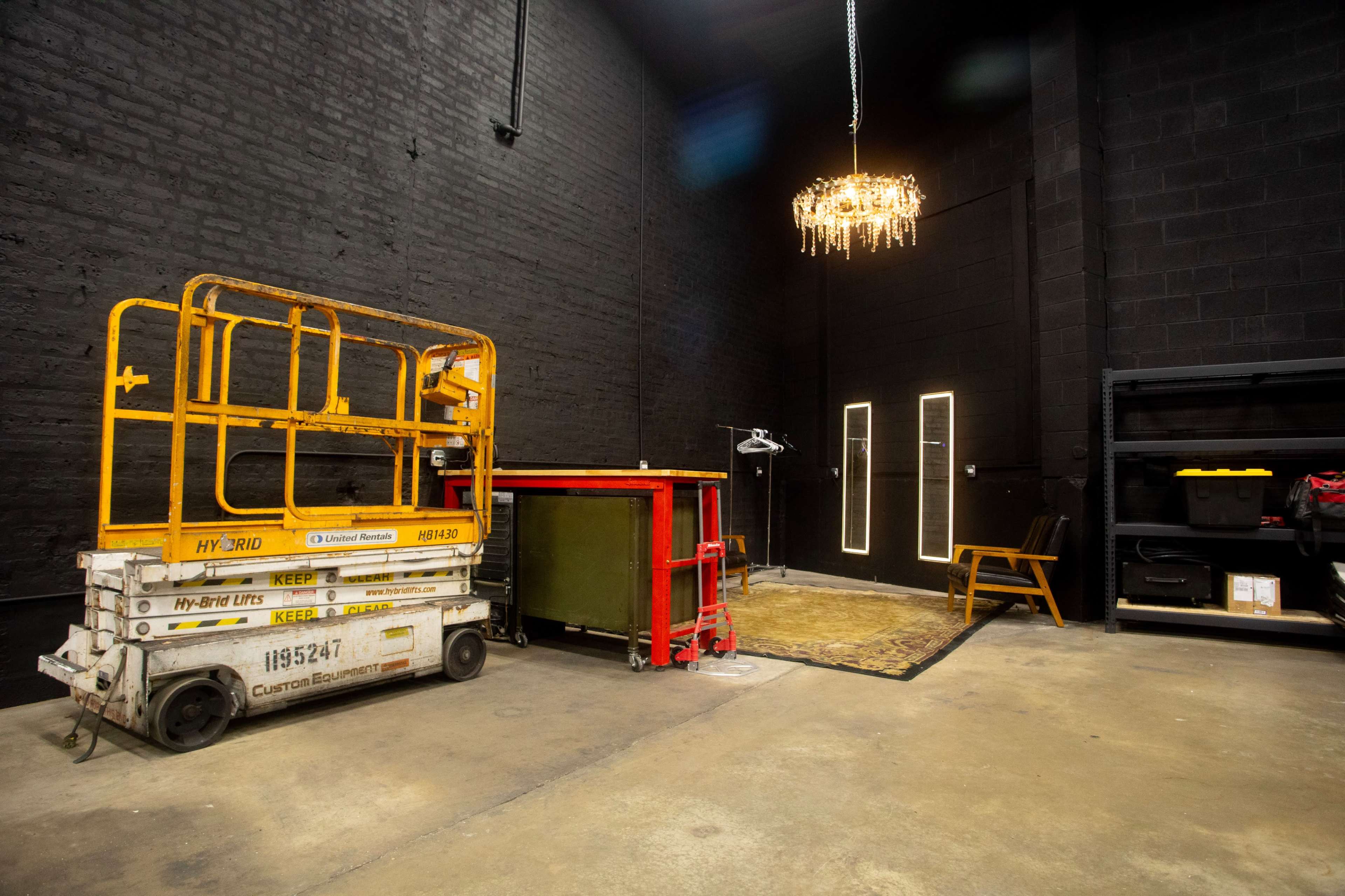 The image shows an empty industrial space with a scissor lift, a few pieces of furniture, a chandelier hanging from the ceiling, and a black wall backdrop.