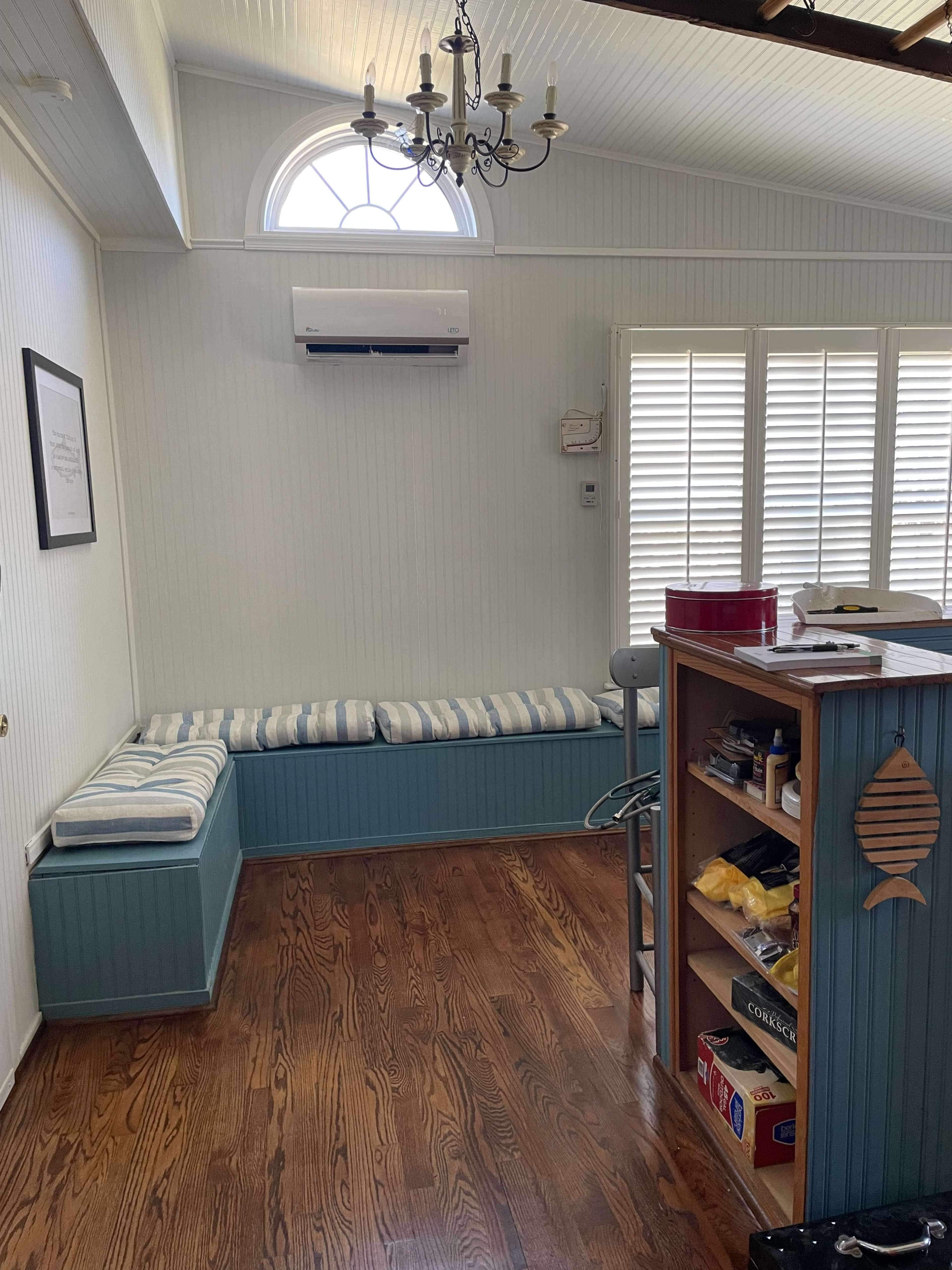 A room with wooden flooring, a blue bench with striped cushions along the wall, and a wooden shelf filled with various items.