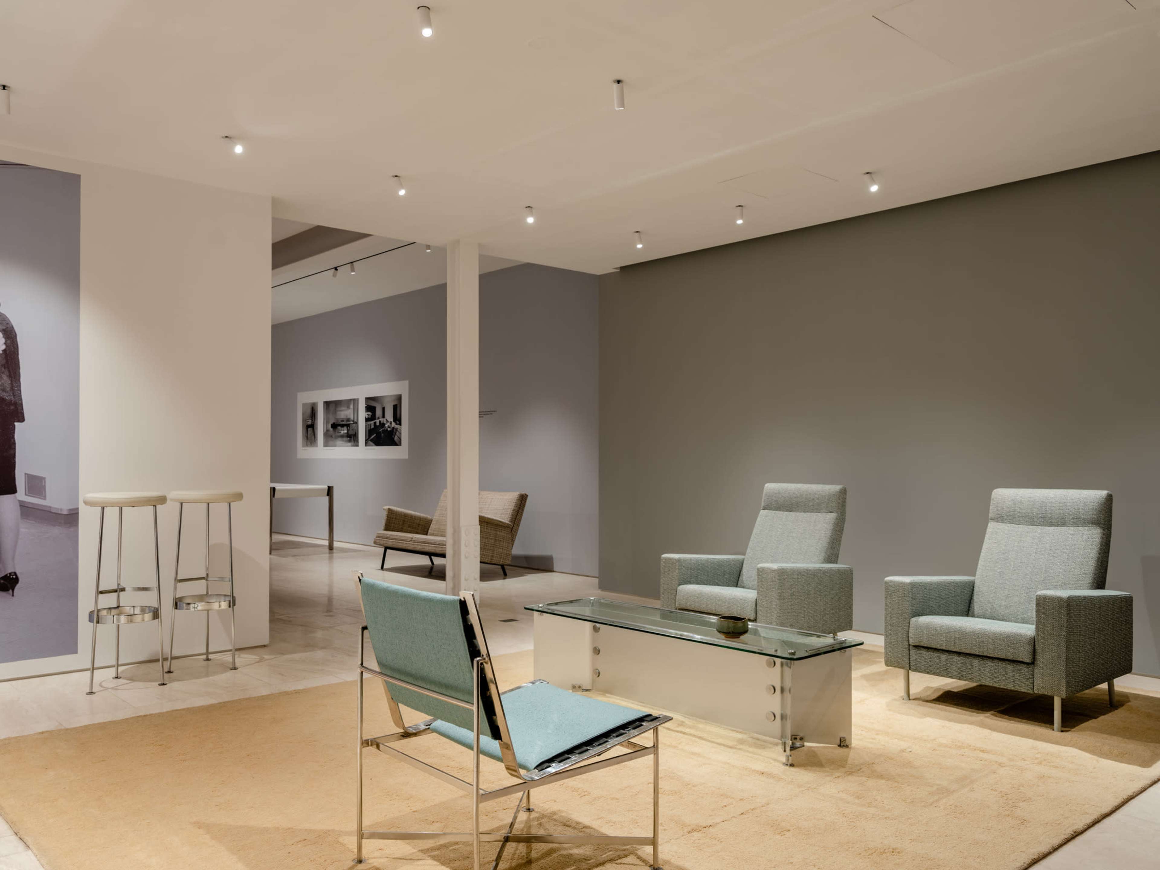 The image shows a modern interior space featuring minimalist seating arrangements, a glass coffee table, and a light-colored area rug.