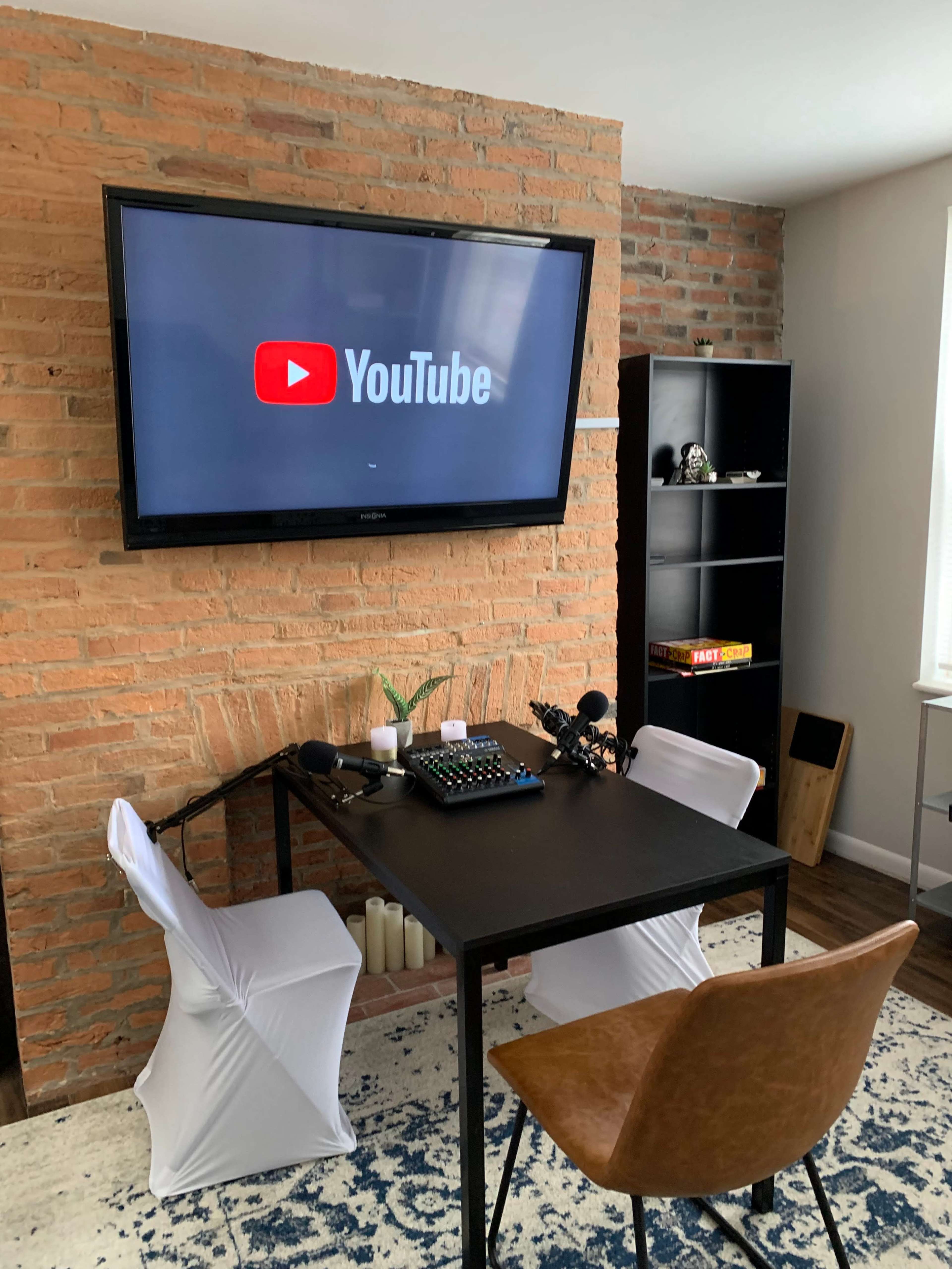 A modern workspace features a table with recording equipment, two chairs, and a wall-mounted TV displaying the YouTube logo against a brick backdrop.