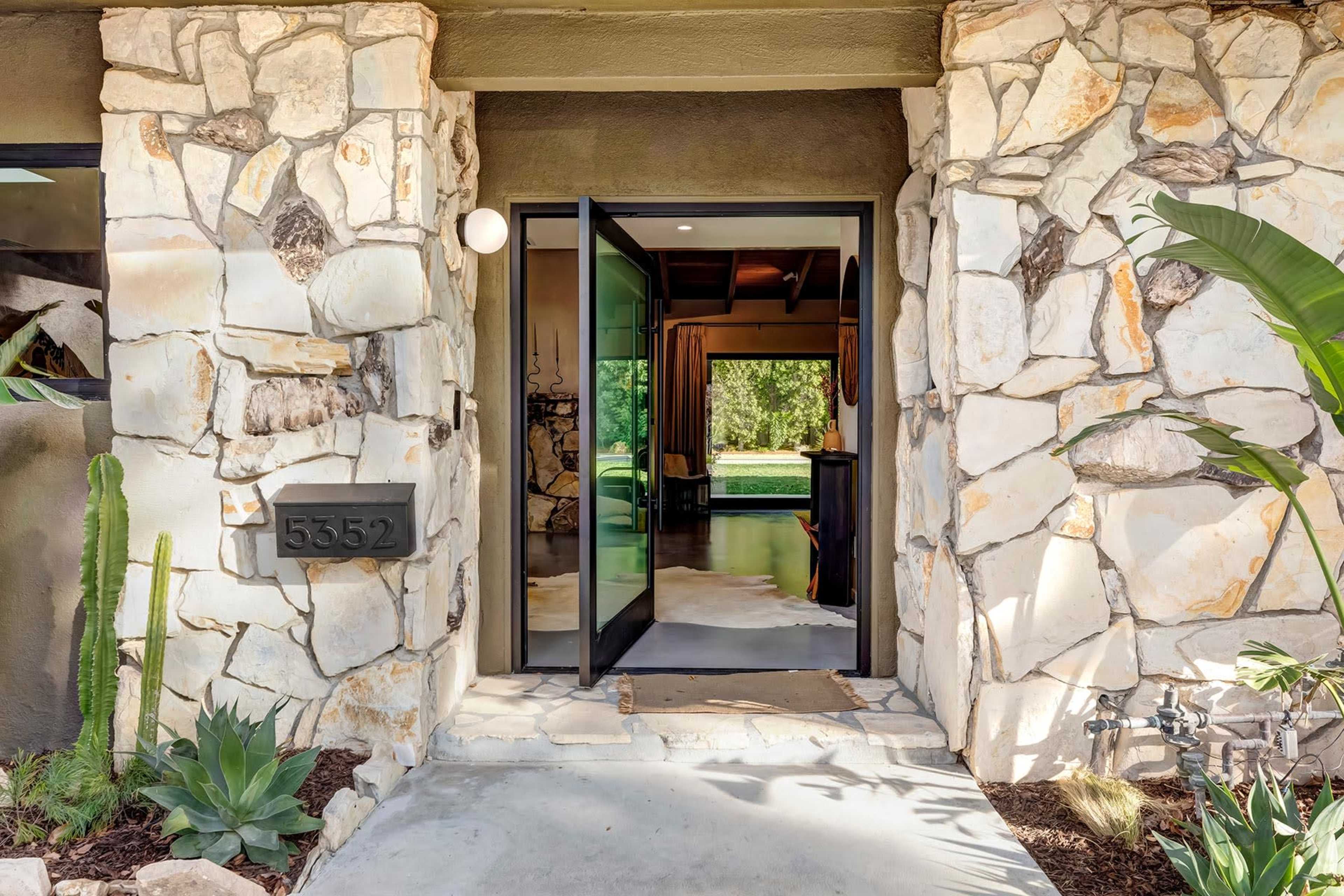 A stone-clad entrance with double glass doors leading into a home, framed by lush plants and a house number displayed on the wall.