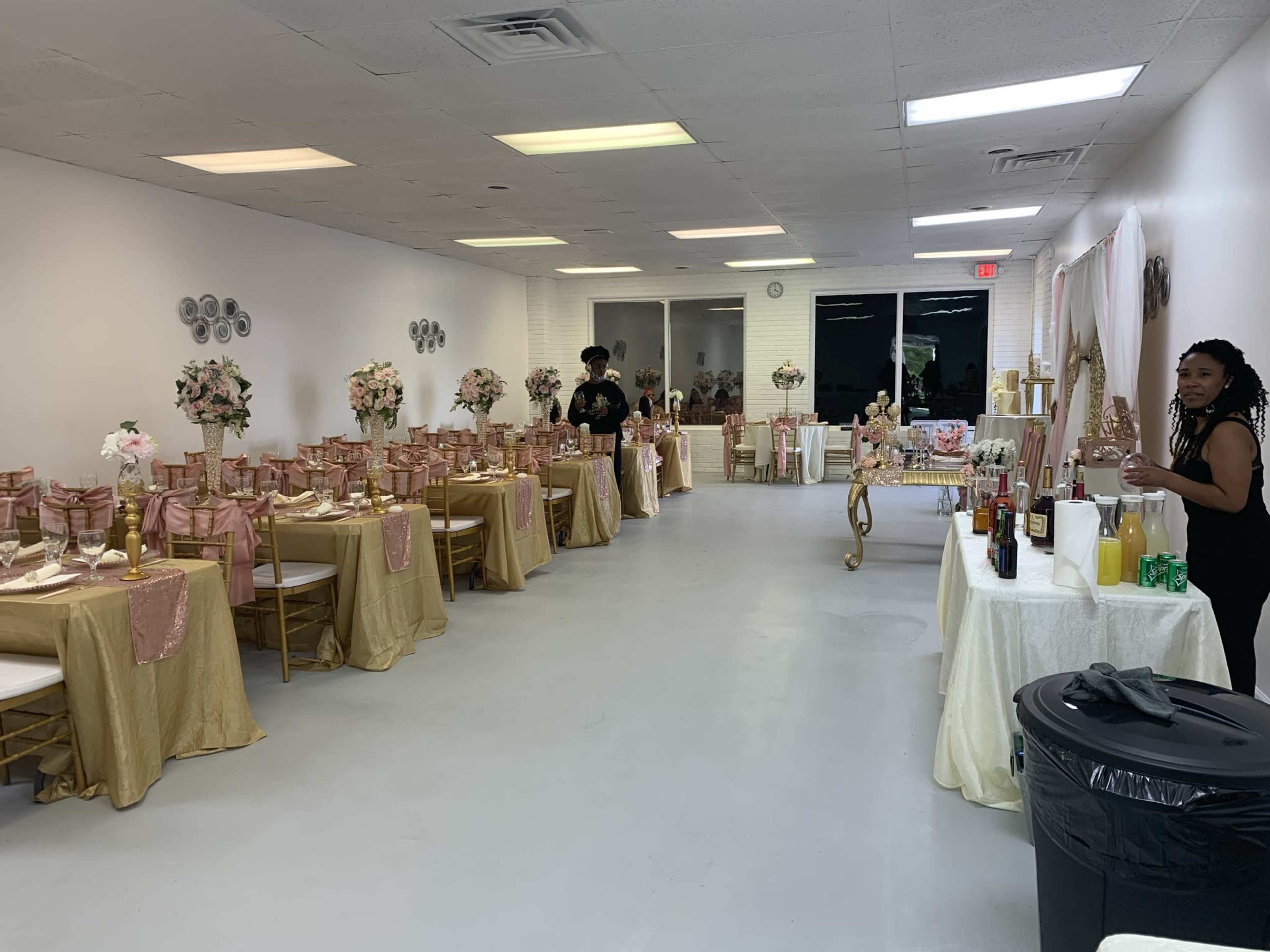 A decorated event space arranged with tables draped in gold and pink tablecloths, floral centerpieces, and a beverage station along one side.