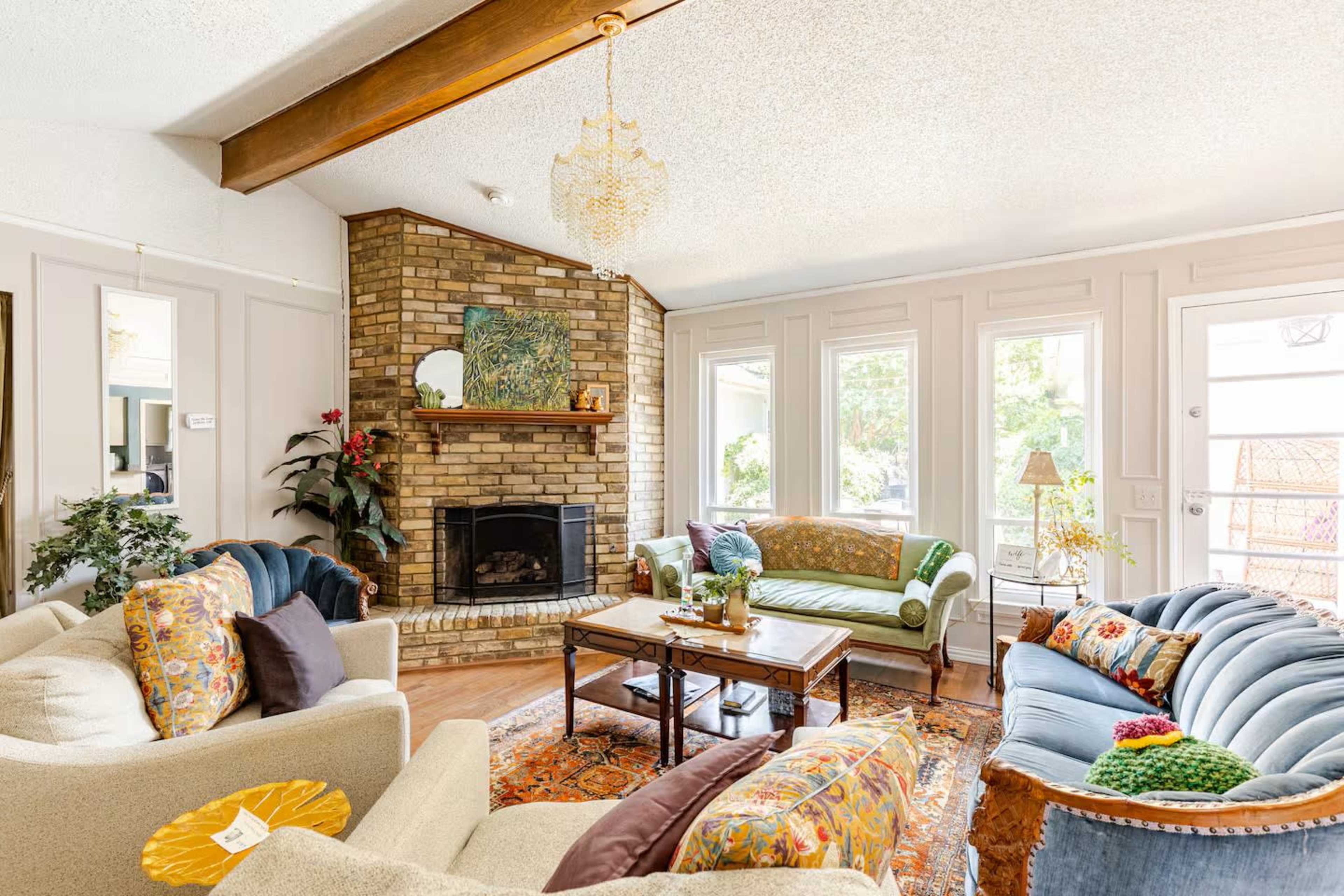 The living room features a brick fireplace, multiple seating arrangements with colorful upholstery, and large windows letting in natural light.