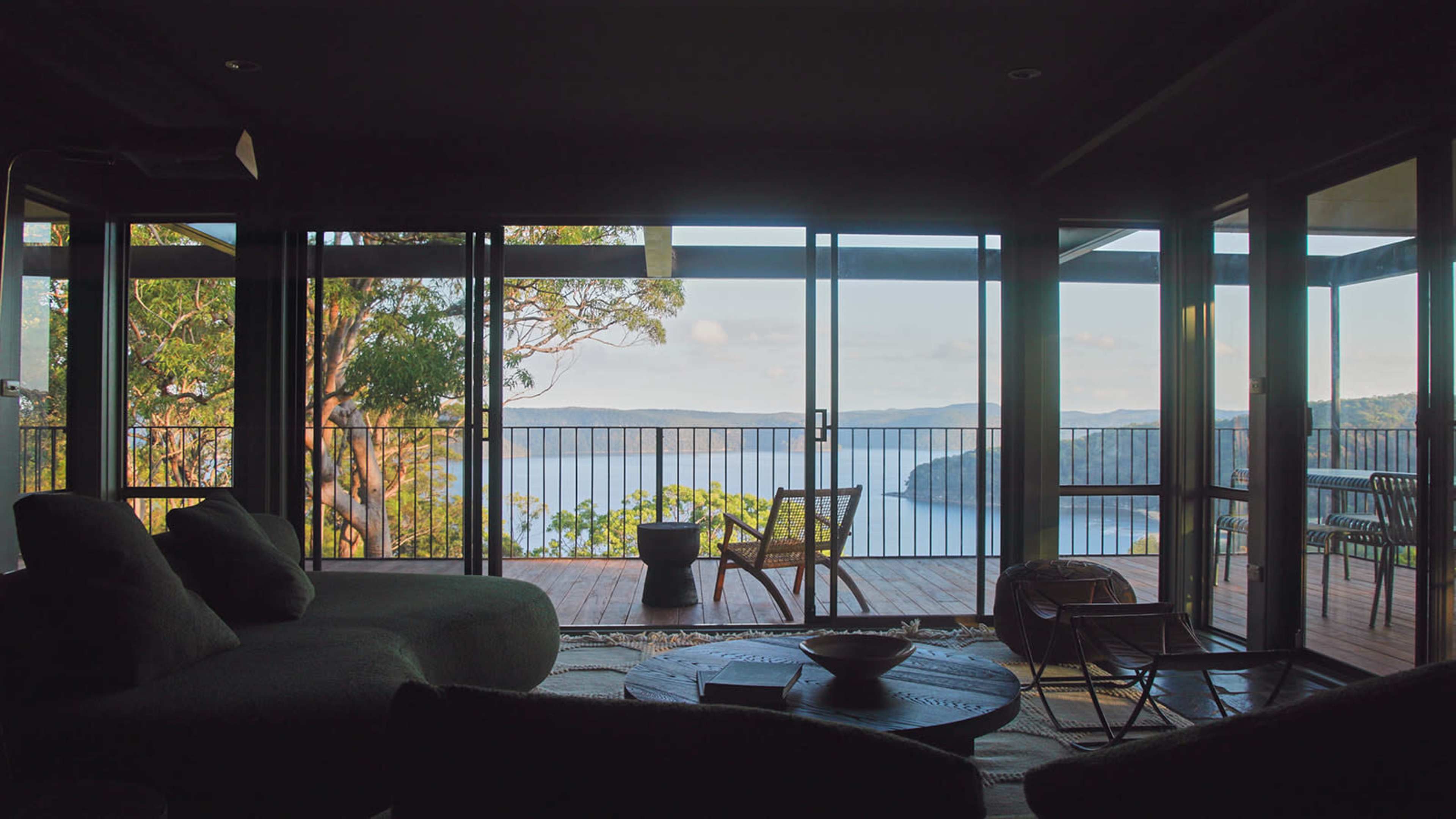 A modern living room features large glass doors opening to a deck with a view of a serene body of water and surrounding landscape.