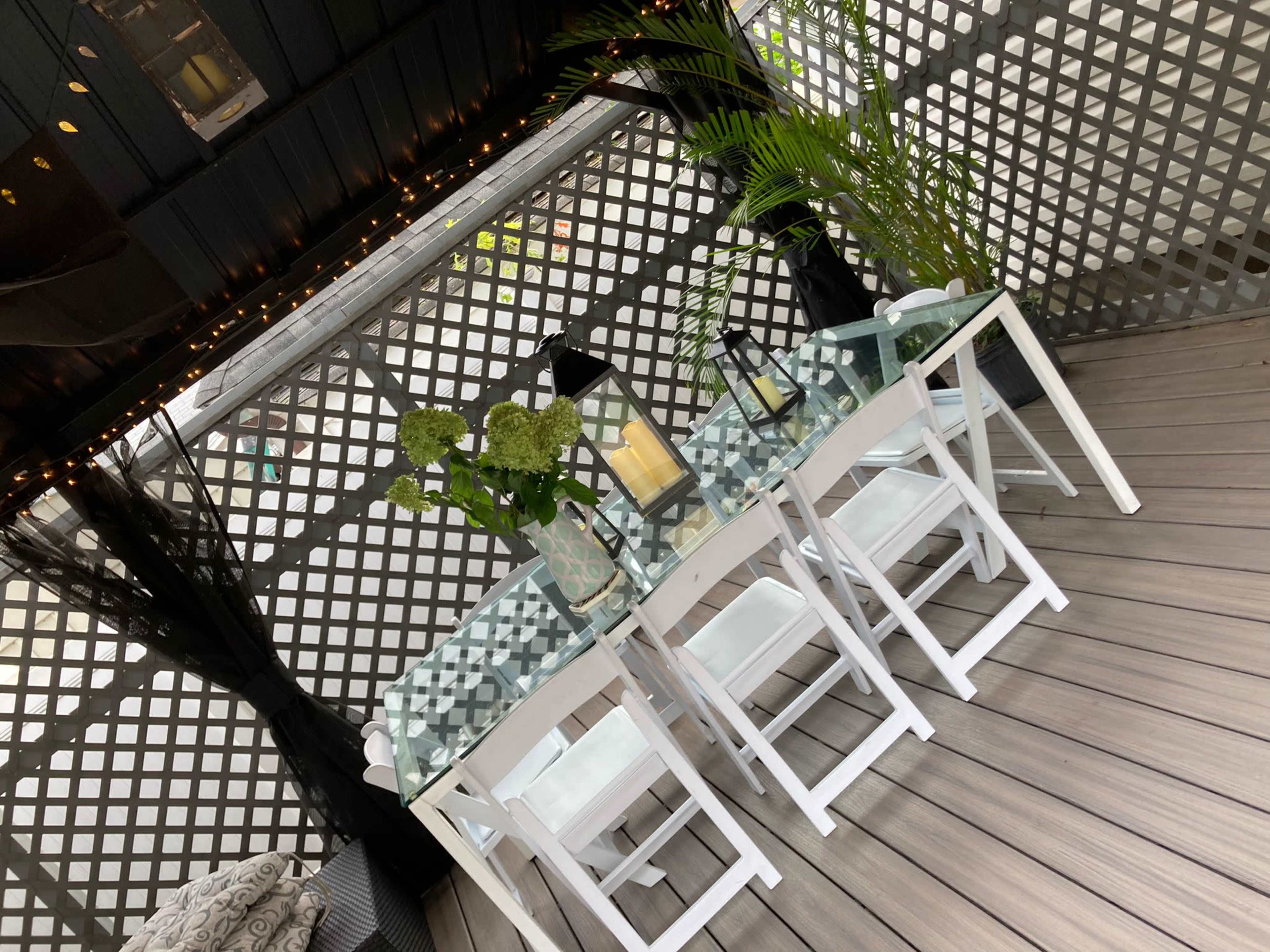 A glass-top dining table with white chairs is arranged under a trellis, surrounded by green plants and soft lighting.