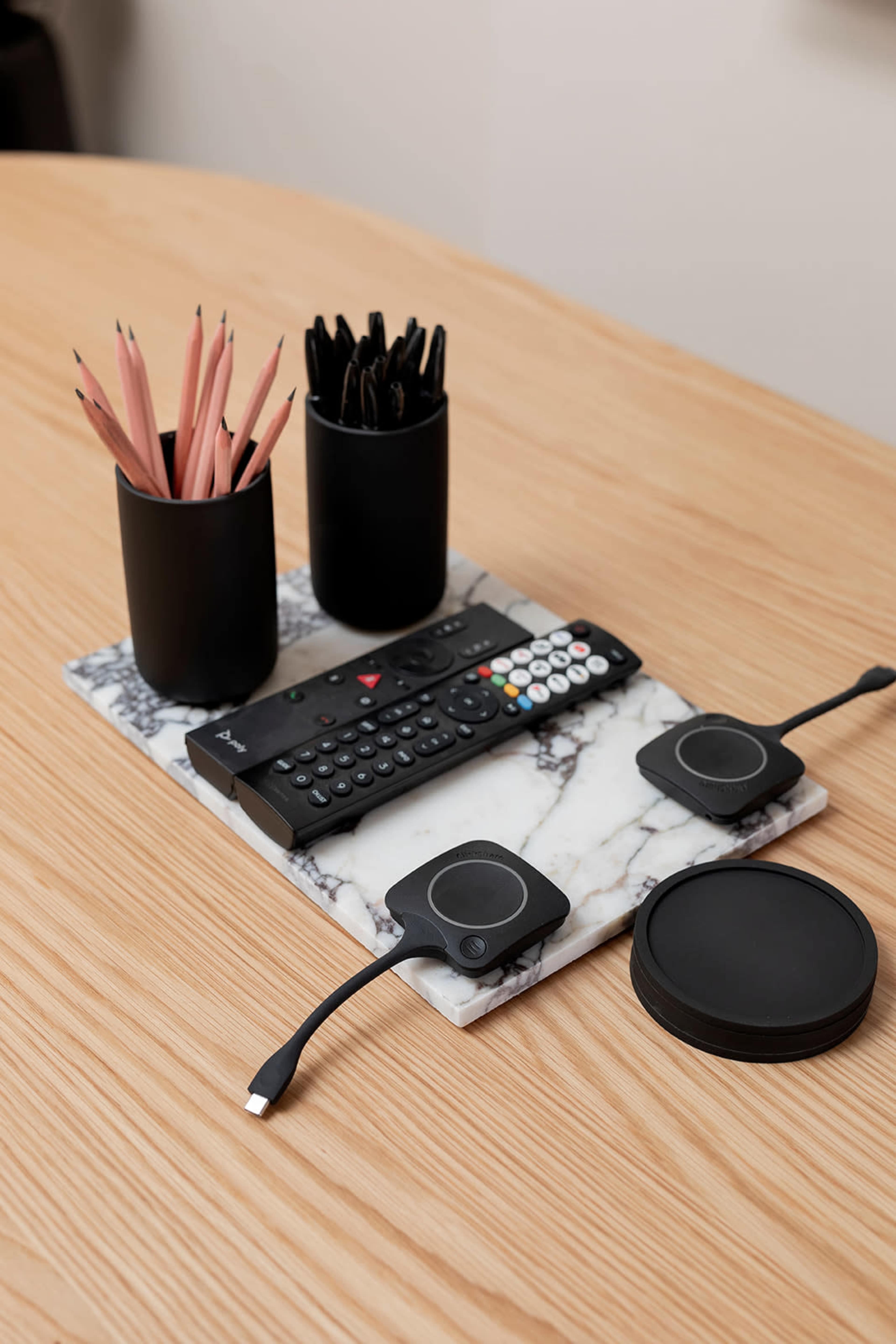 A marble tray on a wooden table holds a remote control, two wireless charging pads, and two black containers filled with pens.