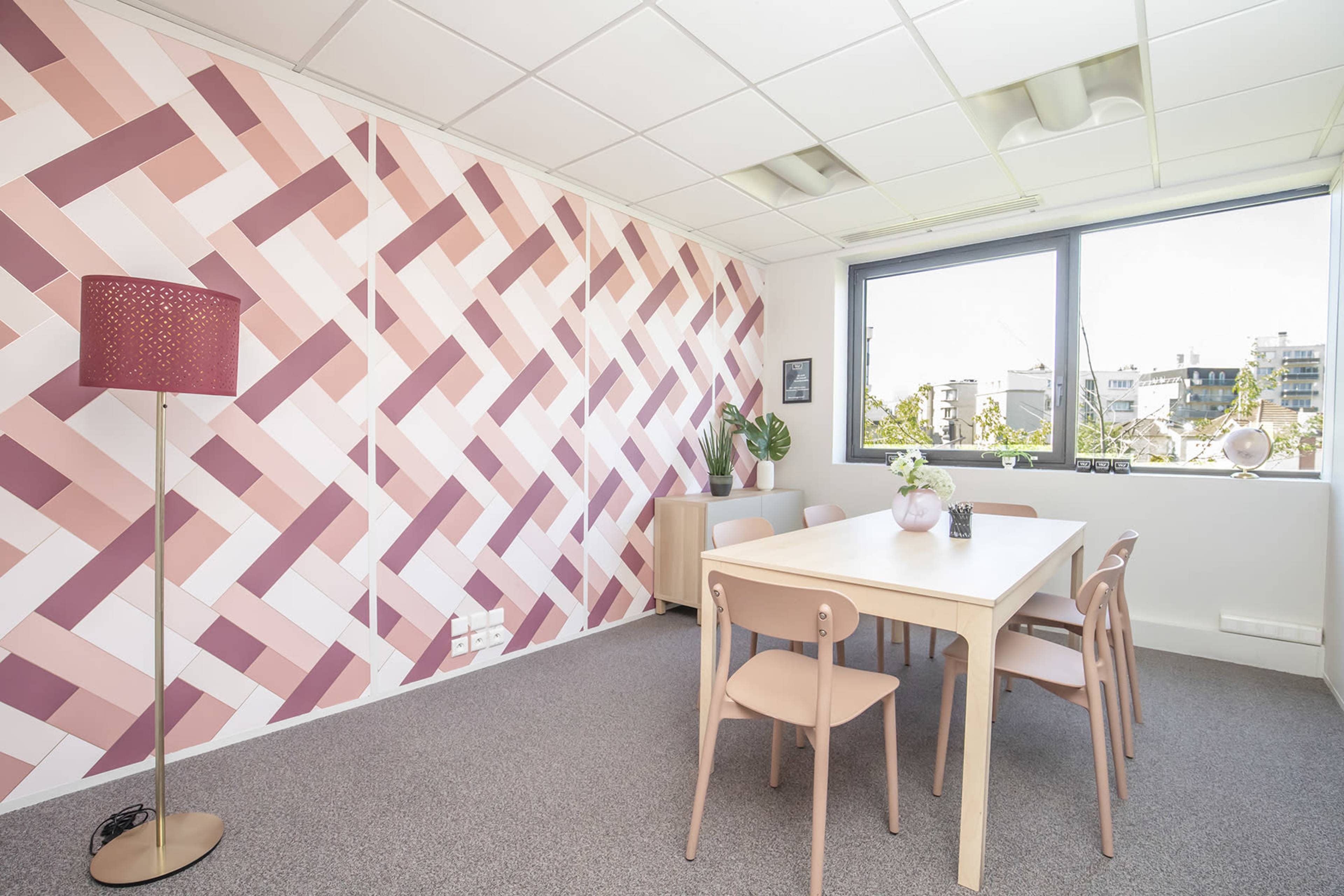 A modern office space featuring a table surrounded by chairs, a potted plant, and a patterned pink and white wall.