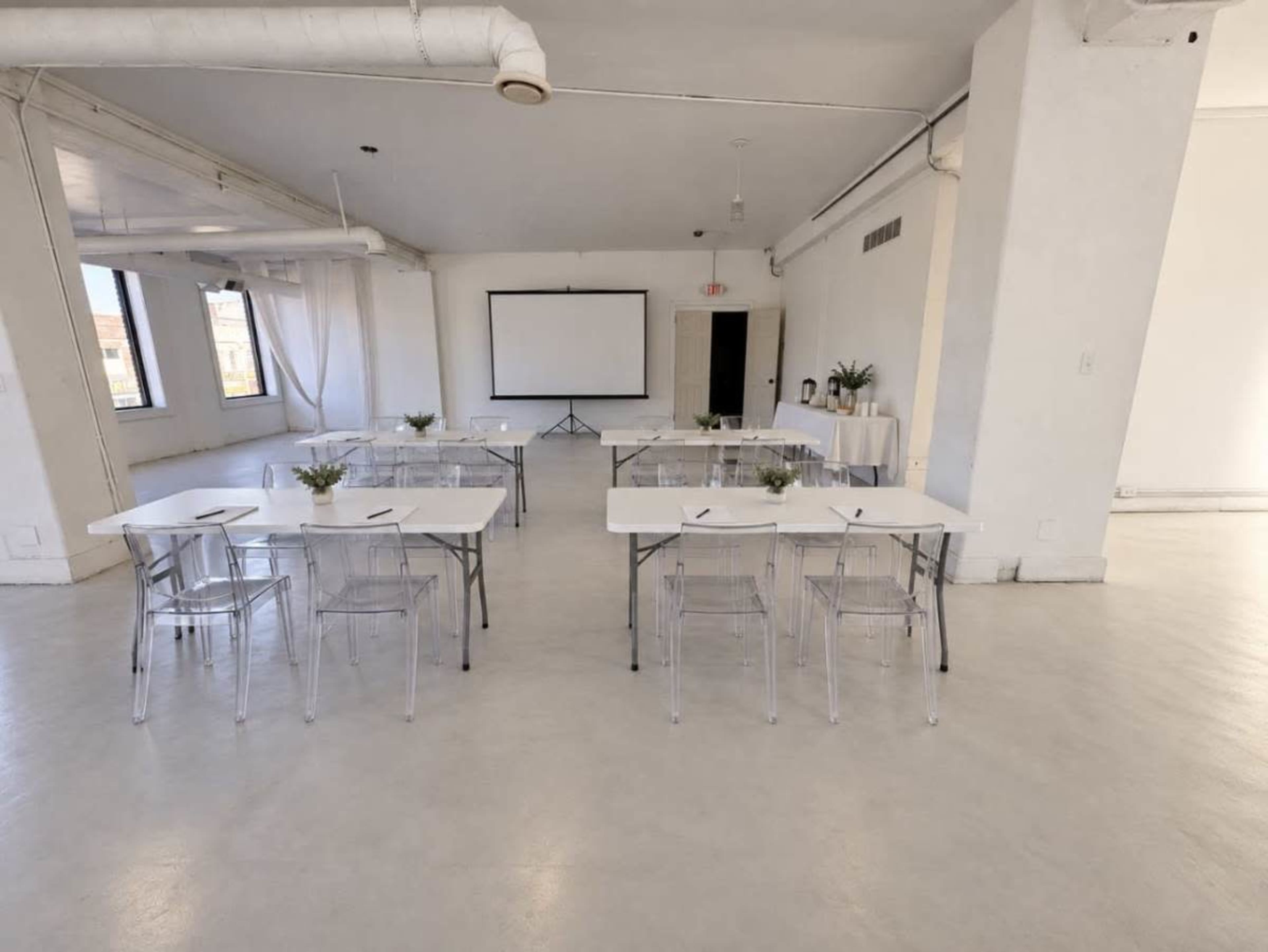A bright, minimalist conference room is set up with several tables and clear chairs, facing a blank presentation screen.