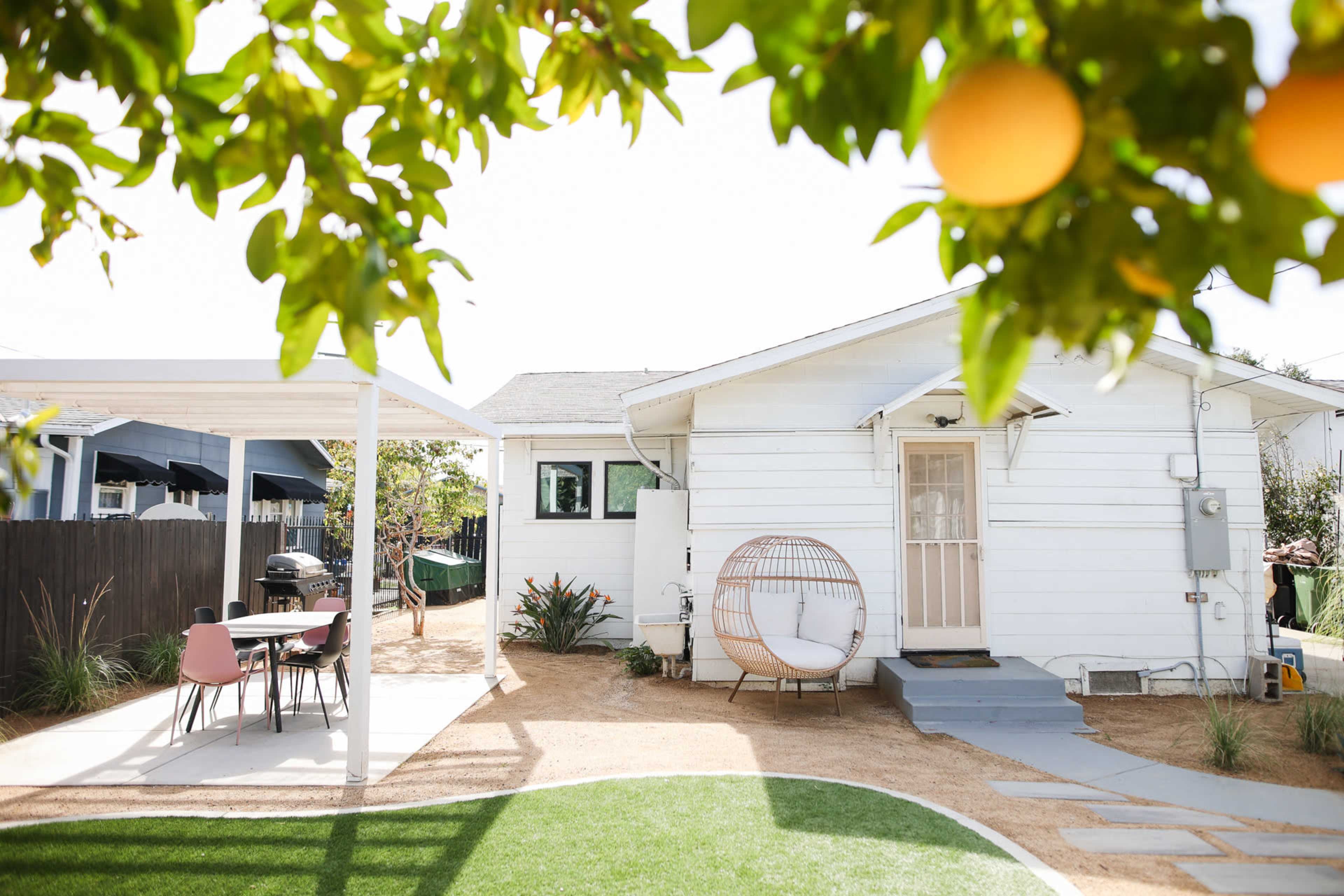A white house with a front yard featuring a circular seating area, a dining table under a pergola, and a lemon tree with ripe lemons.