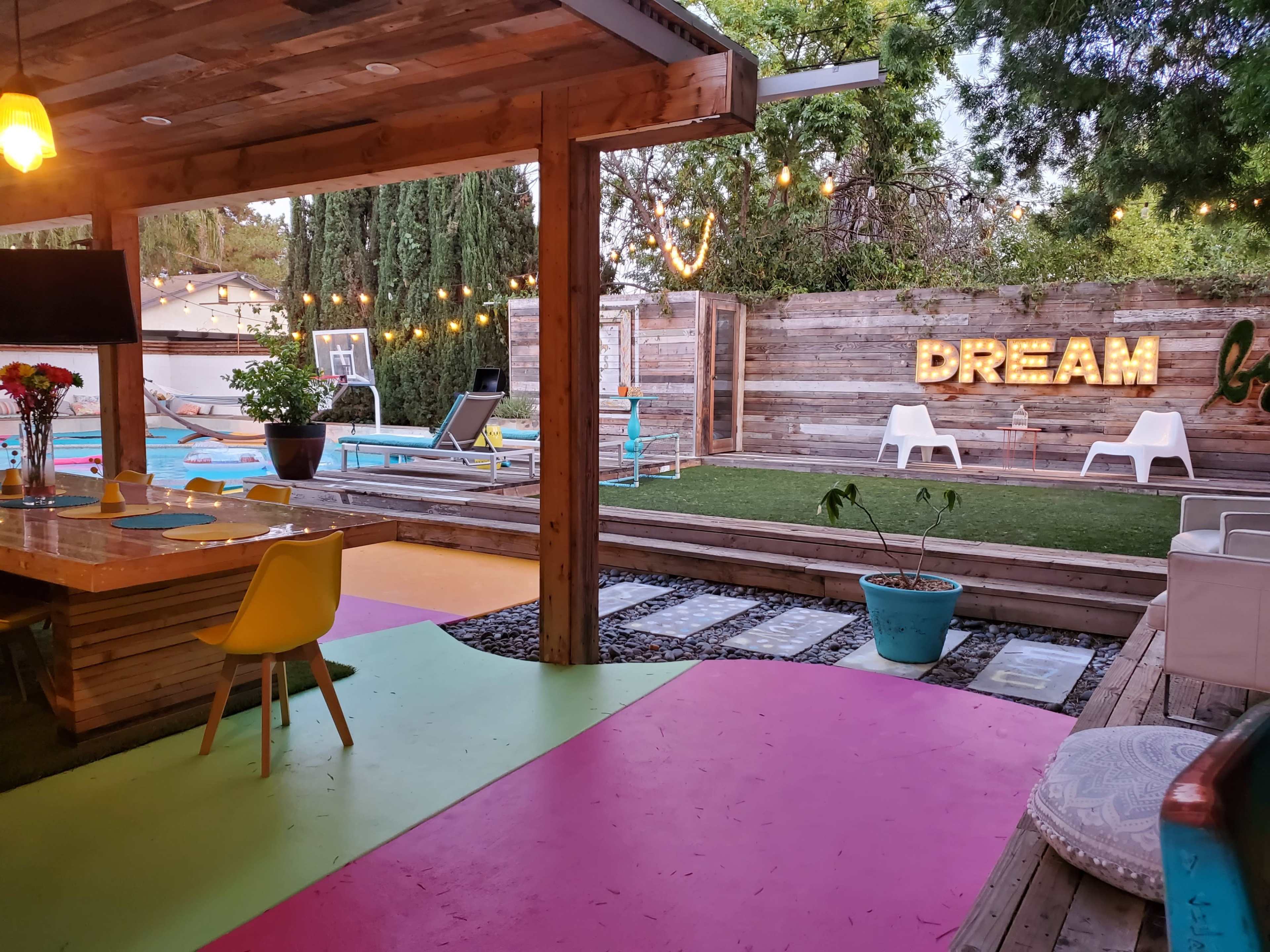 The image shows a colorful outdoor area with a dining table, lounge chairs, a pool, and a lit sign displaying the word "DREAM" against a wooden backdrop.