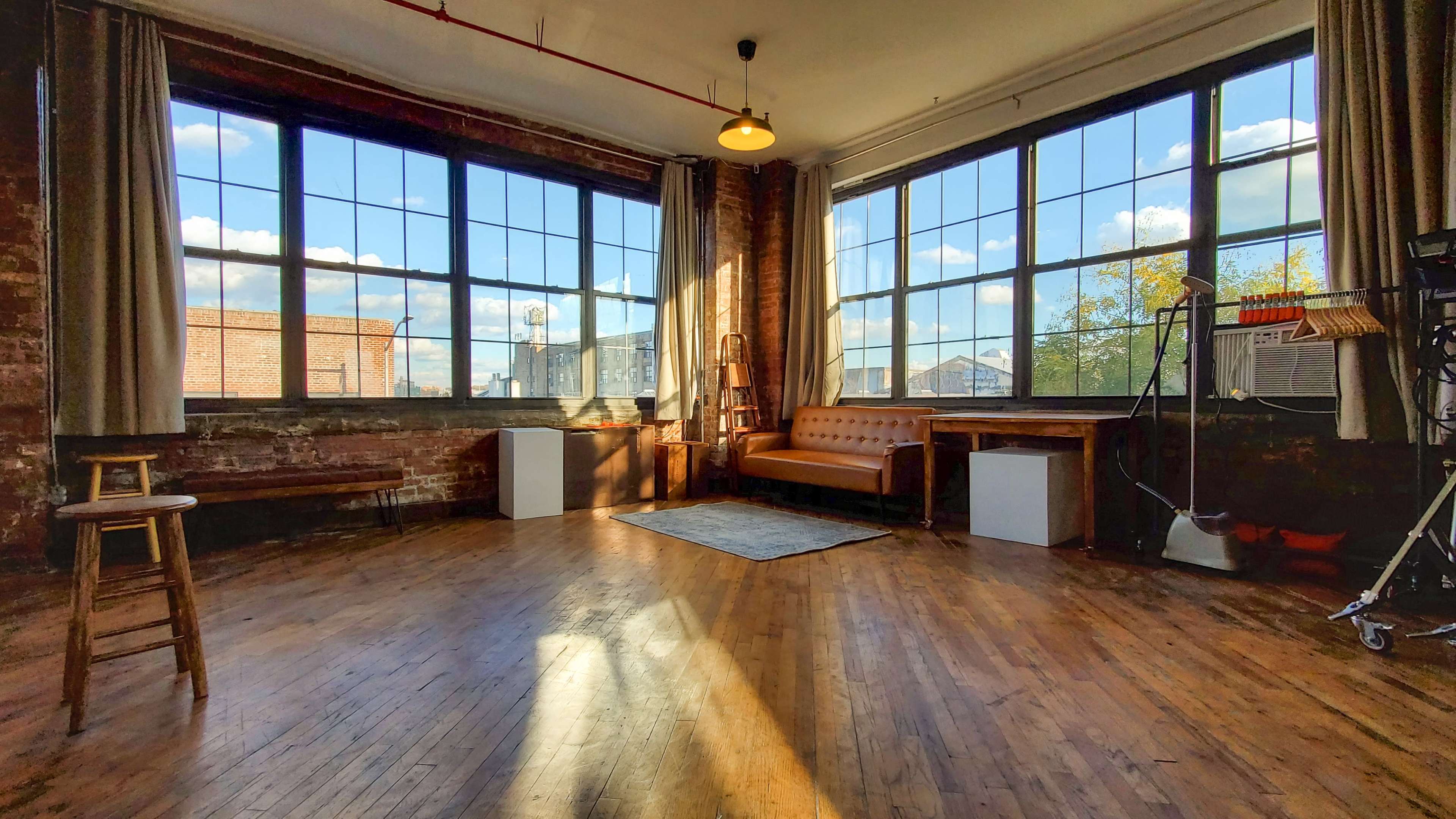 A spacious room with large windows, wooden flooring, a sofa, a table, and a ladder against a brick wall.