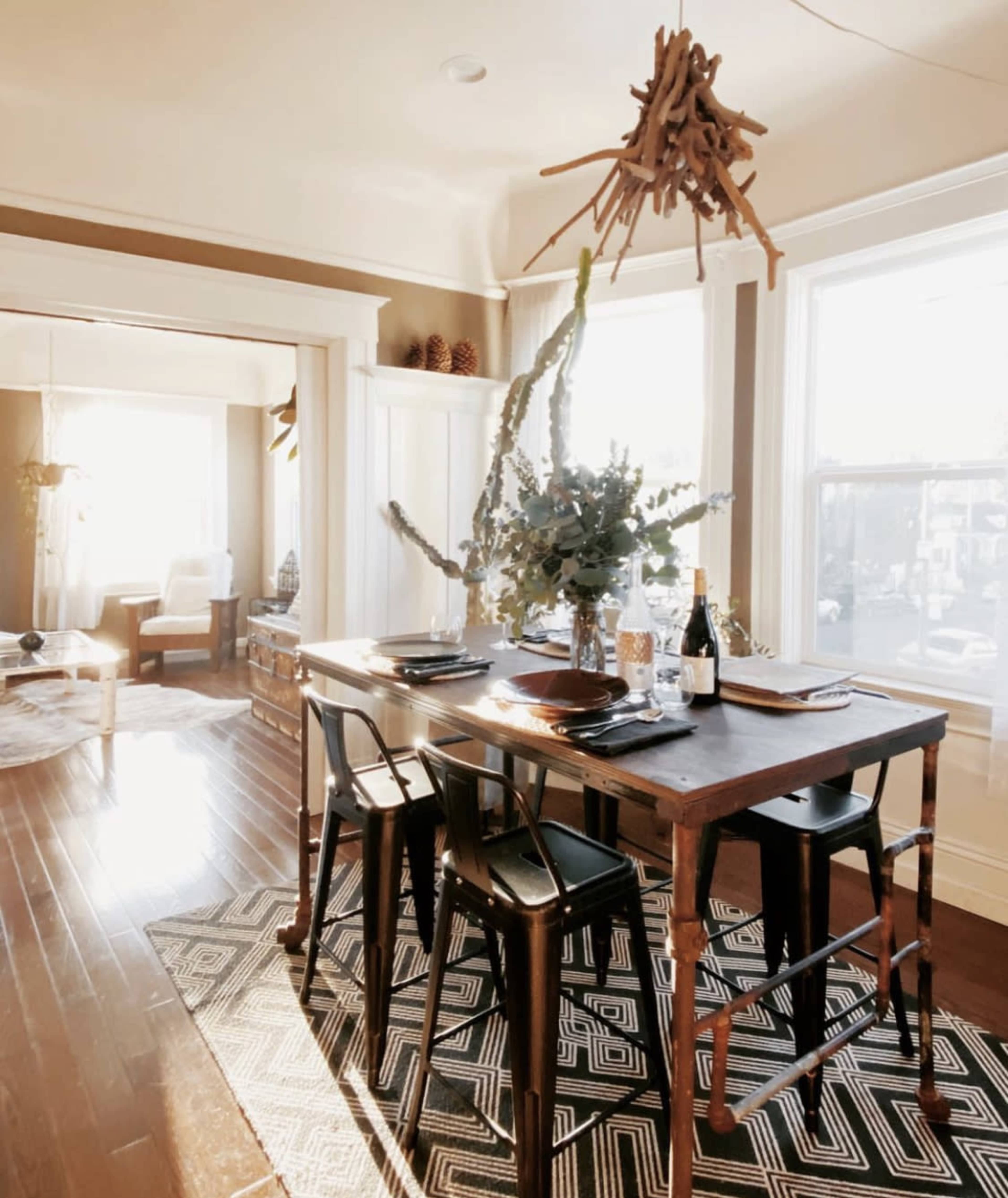 A wooden dining table is set with dishes and a bottle of champagne, surrounded by chairs and decorated with a tall arrangement of branches, all illuminated by natural light through large windows.