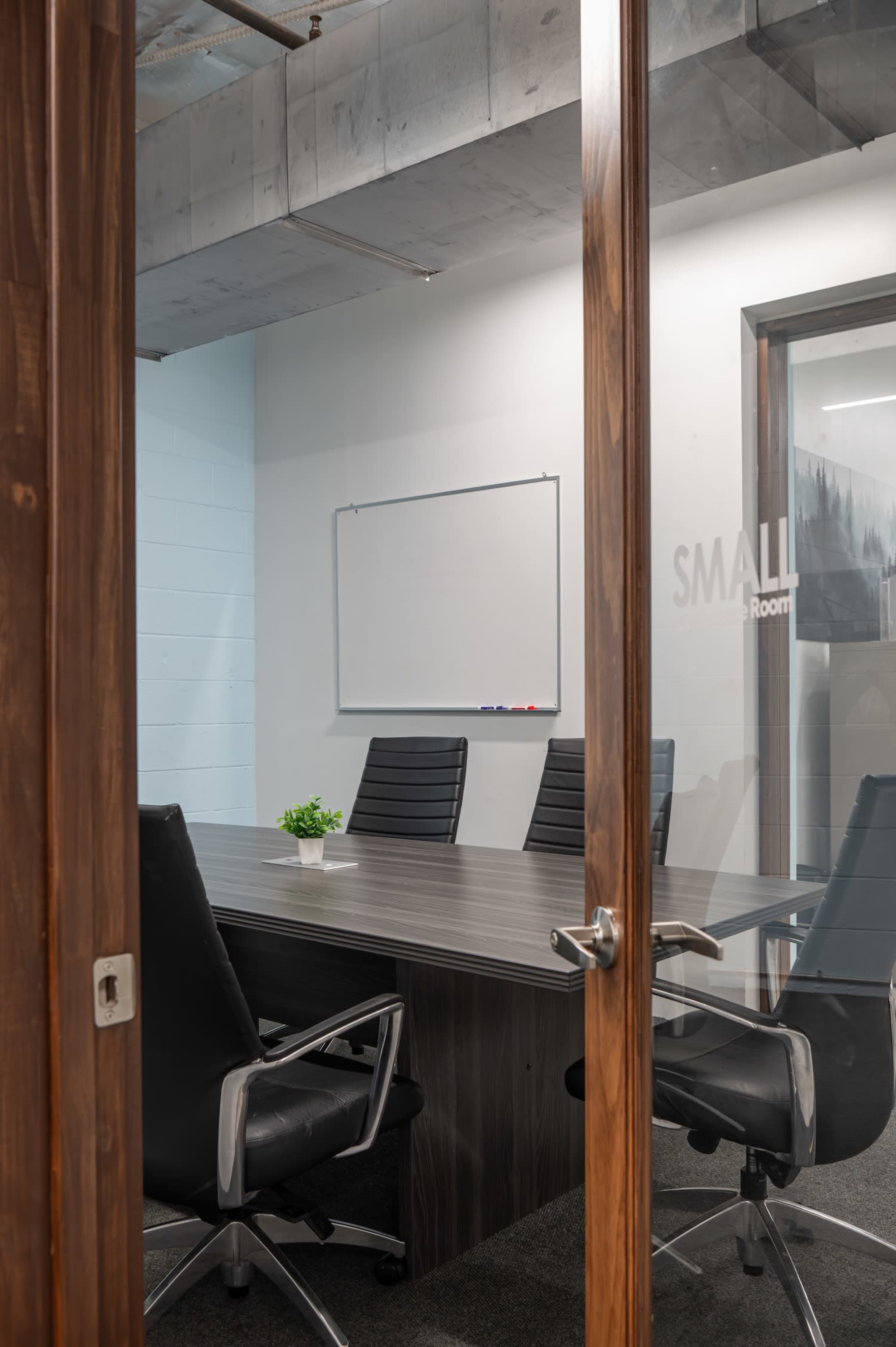 The image shows a modern conference room featuring a large wooden table surrounded by black leather chairs, with a whiteboard on the wall and a small plant on the table.