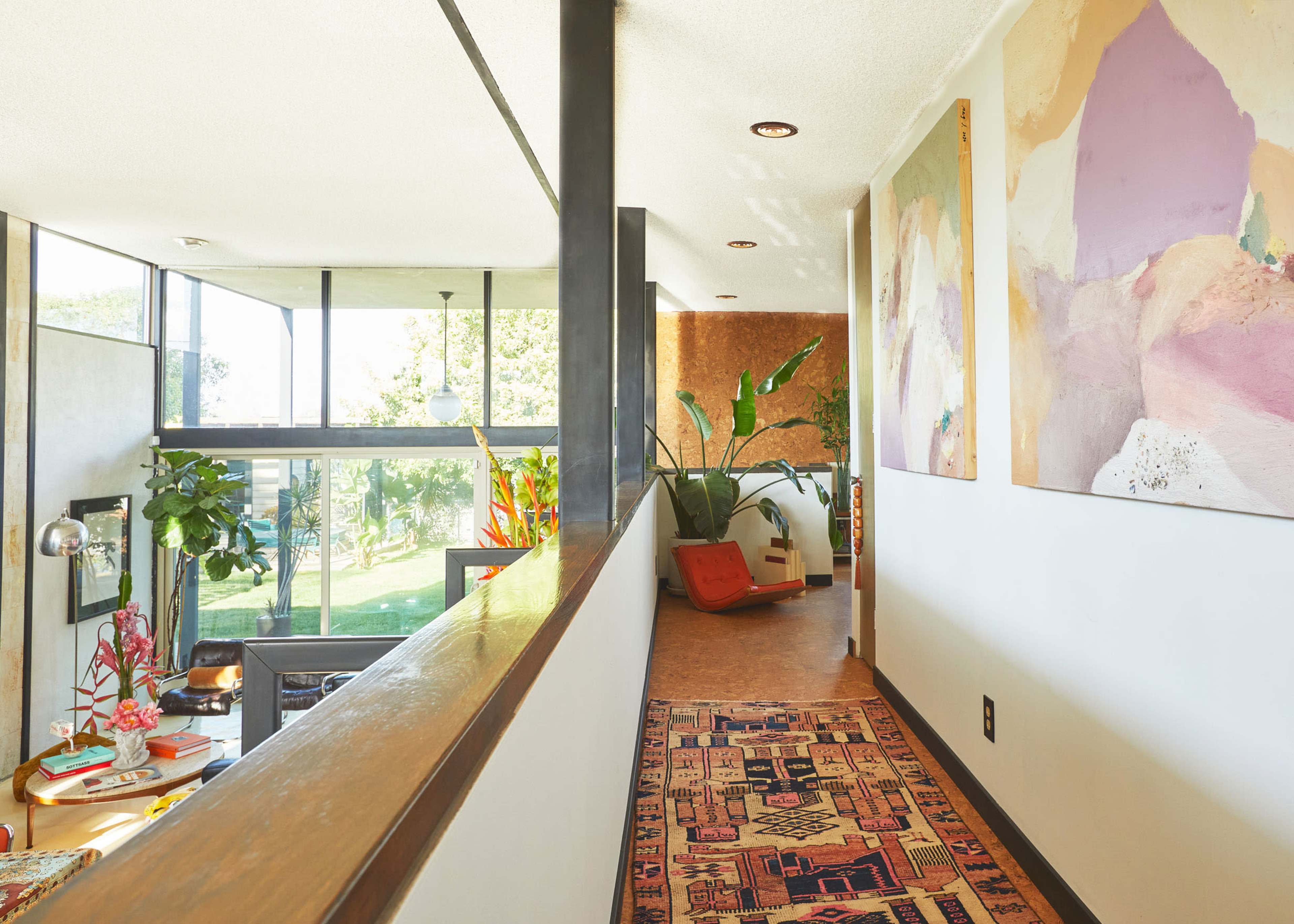 A hallway lined with artwork leads to a living space featuring large windows and decorative plants.