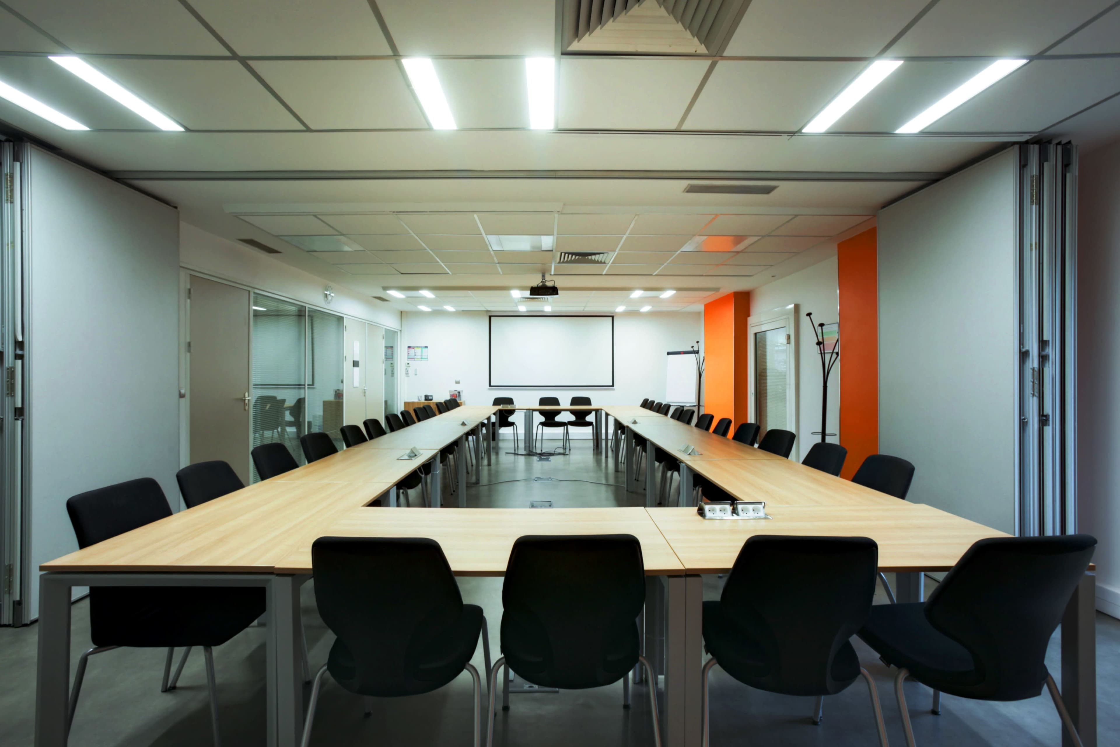 A large, modern conference room features a long table surrounded by black chairs, with a projector screen at one end and orange accent walls.