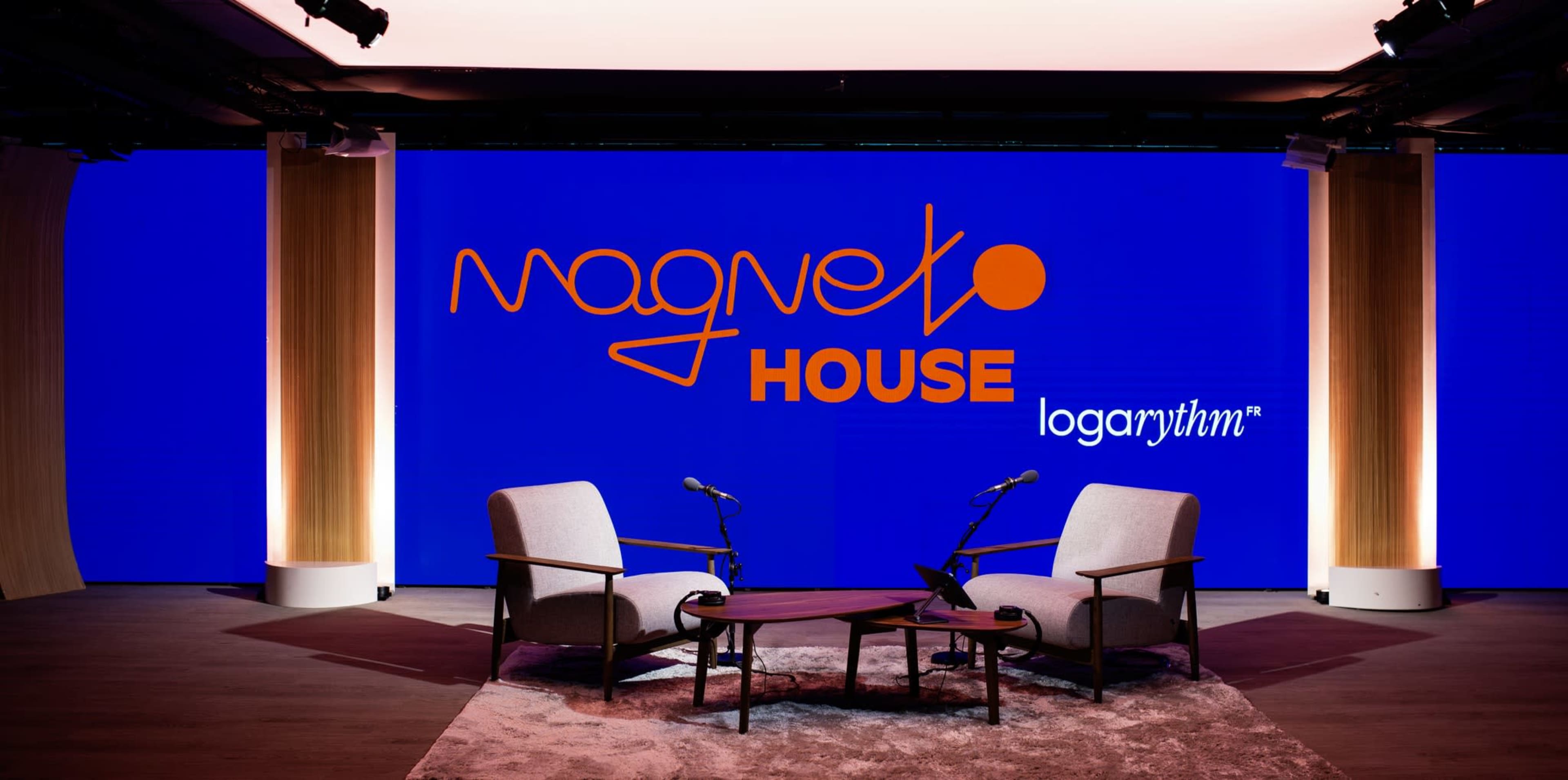 The image shows a modern stage setup with two armchairs facing each other in front of a large lit backdrop displaying the text "Magneto House" and "logarithm."