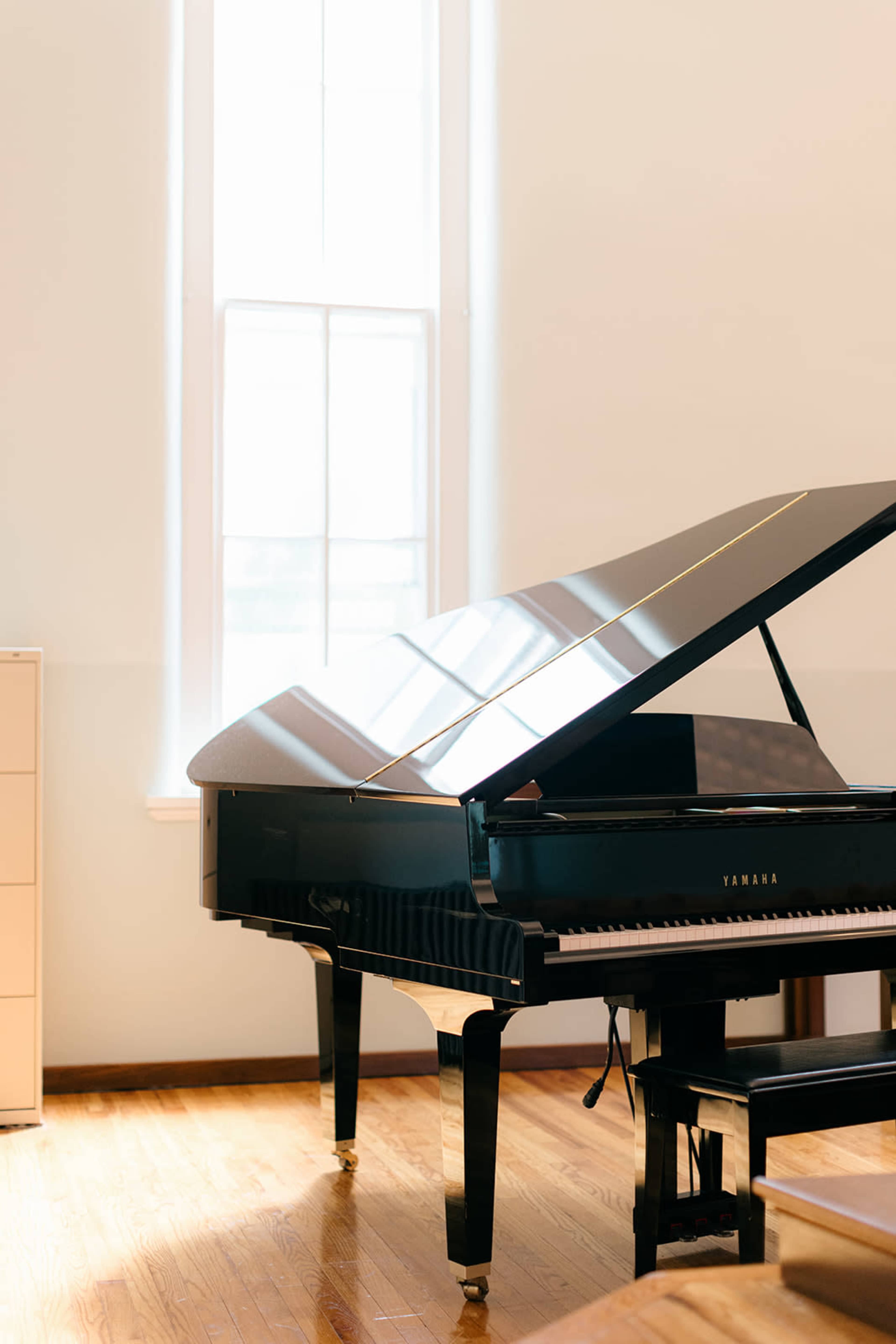 A grand piano is positioned in a well-lit room beside a tall window, with light reflecting off its polished surface.