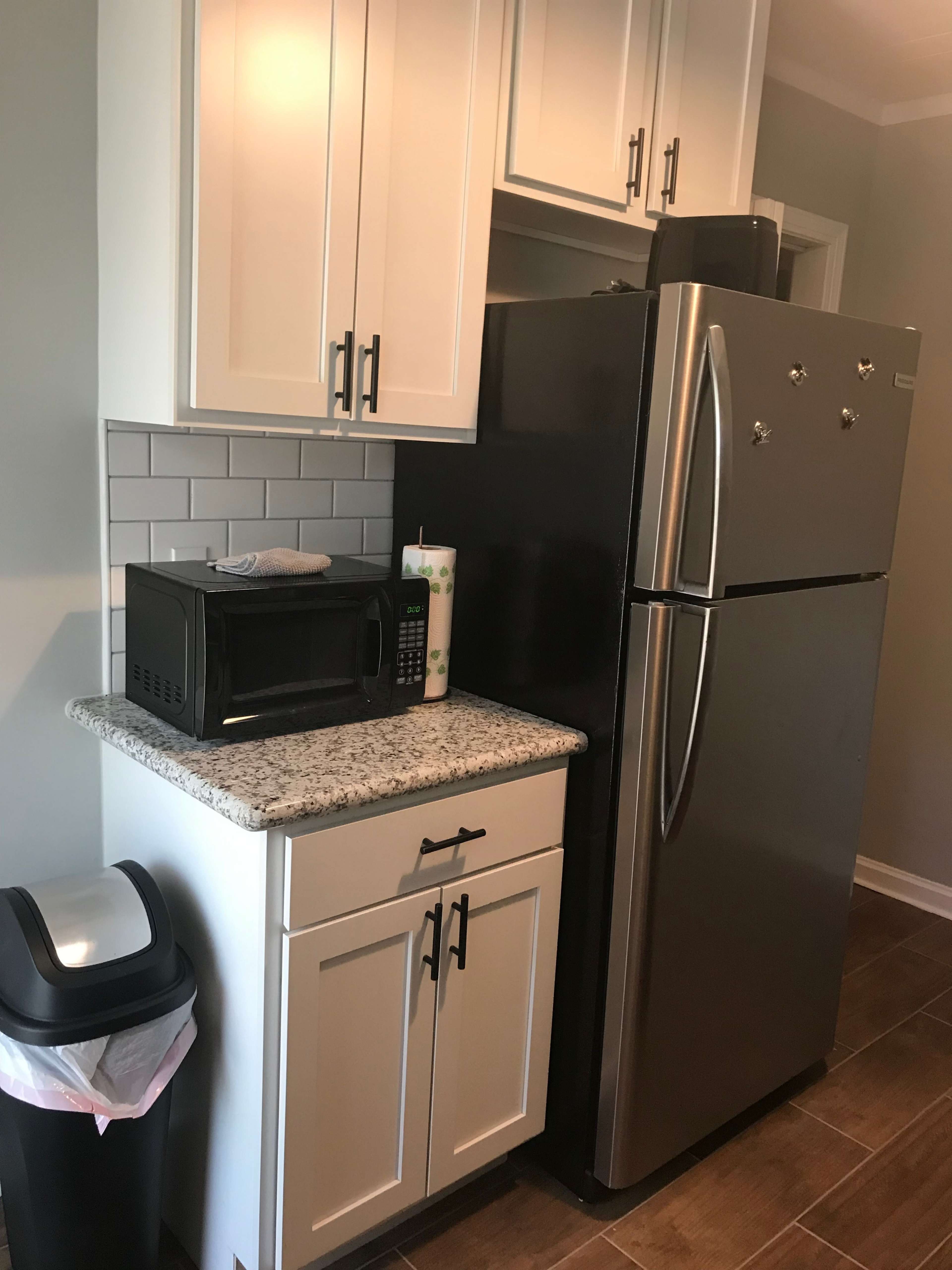 A kitchen corner featuring a granite countertop, a black microwave, a stainless steel refrigerator, and a trash can.