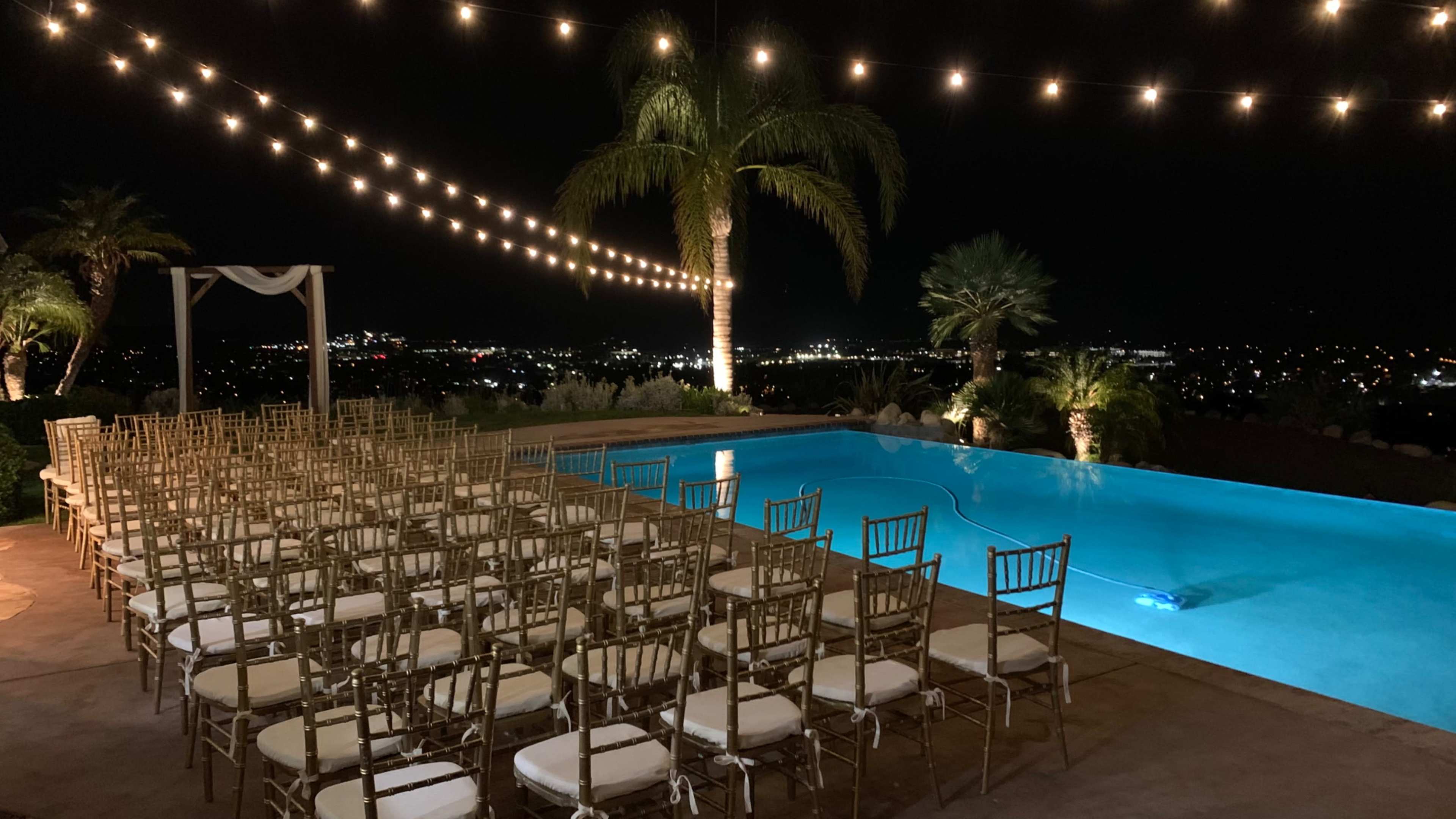 The scene features an outdoor event space at night, with rows of gold chiavari chairs facing a brightly lit pool and string lights overhead, surrounded by palm trees and a view of city lights in the distance.