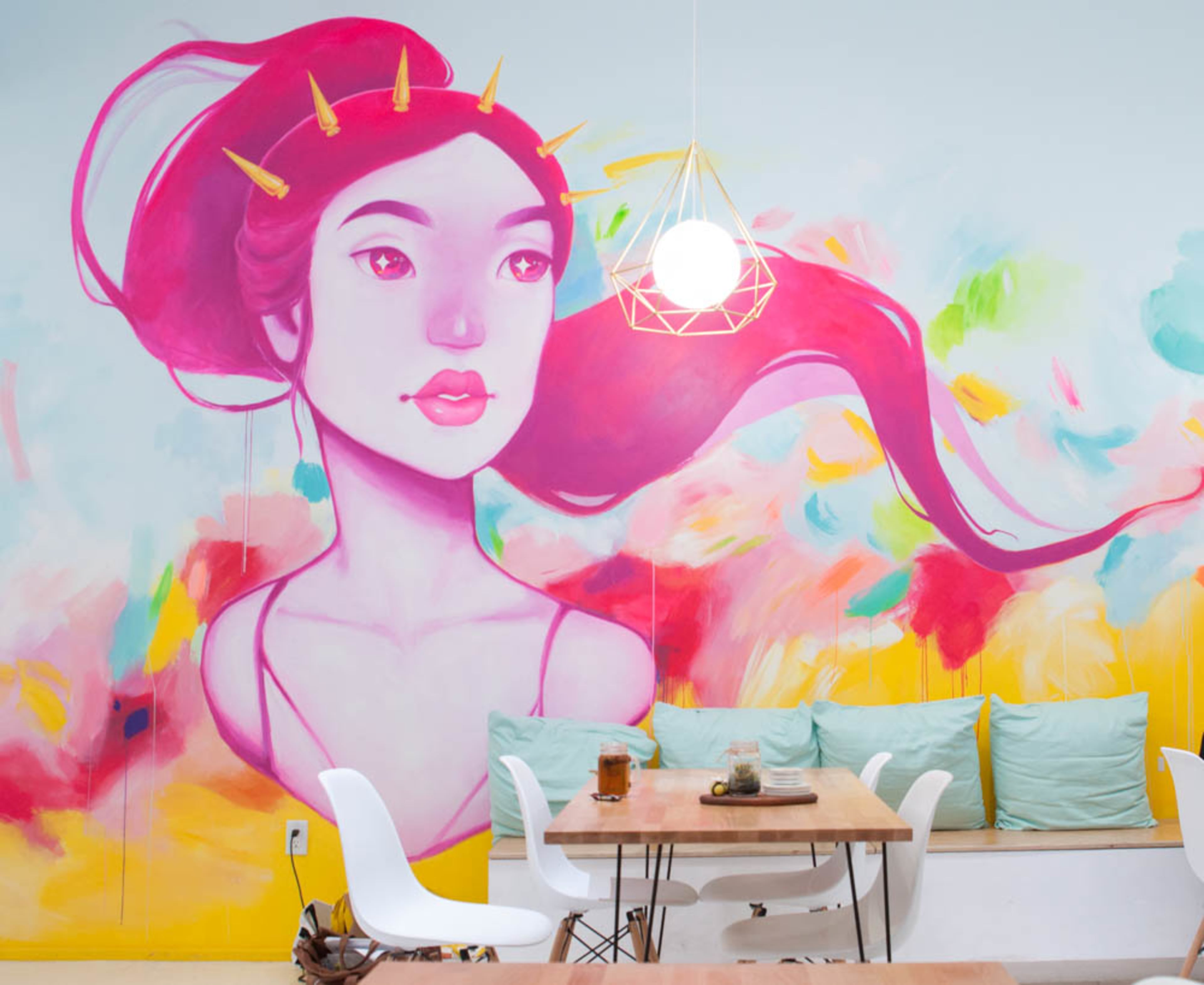 A vibrant mural of a woman with long, flowing hair and decorative hairpins, set against a colorful background in a café with light wood tables and white chairs.