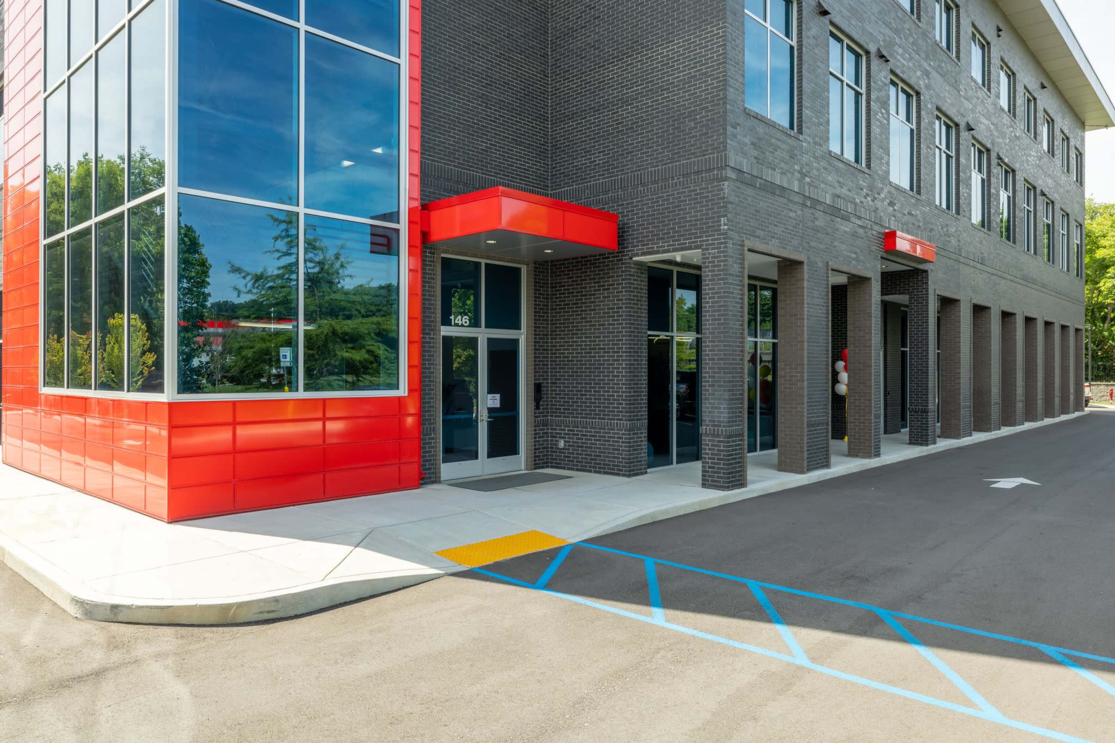 A modern building with a red and gray exterior, featuring large glass windows and a concrete pathway leading to the entrance.