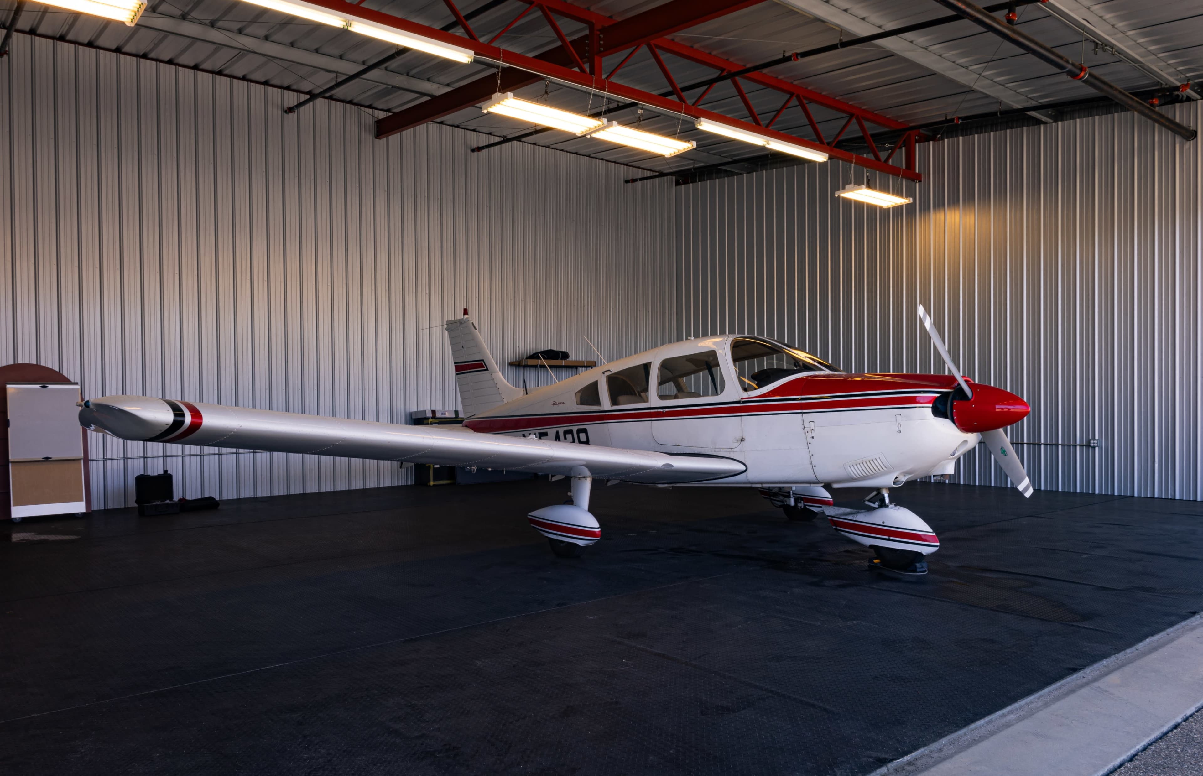 A single-engine airplane with a red and white design is parked inside a hangar with metal walls and a black floor.