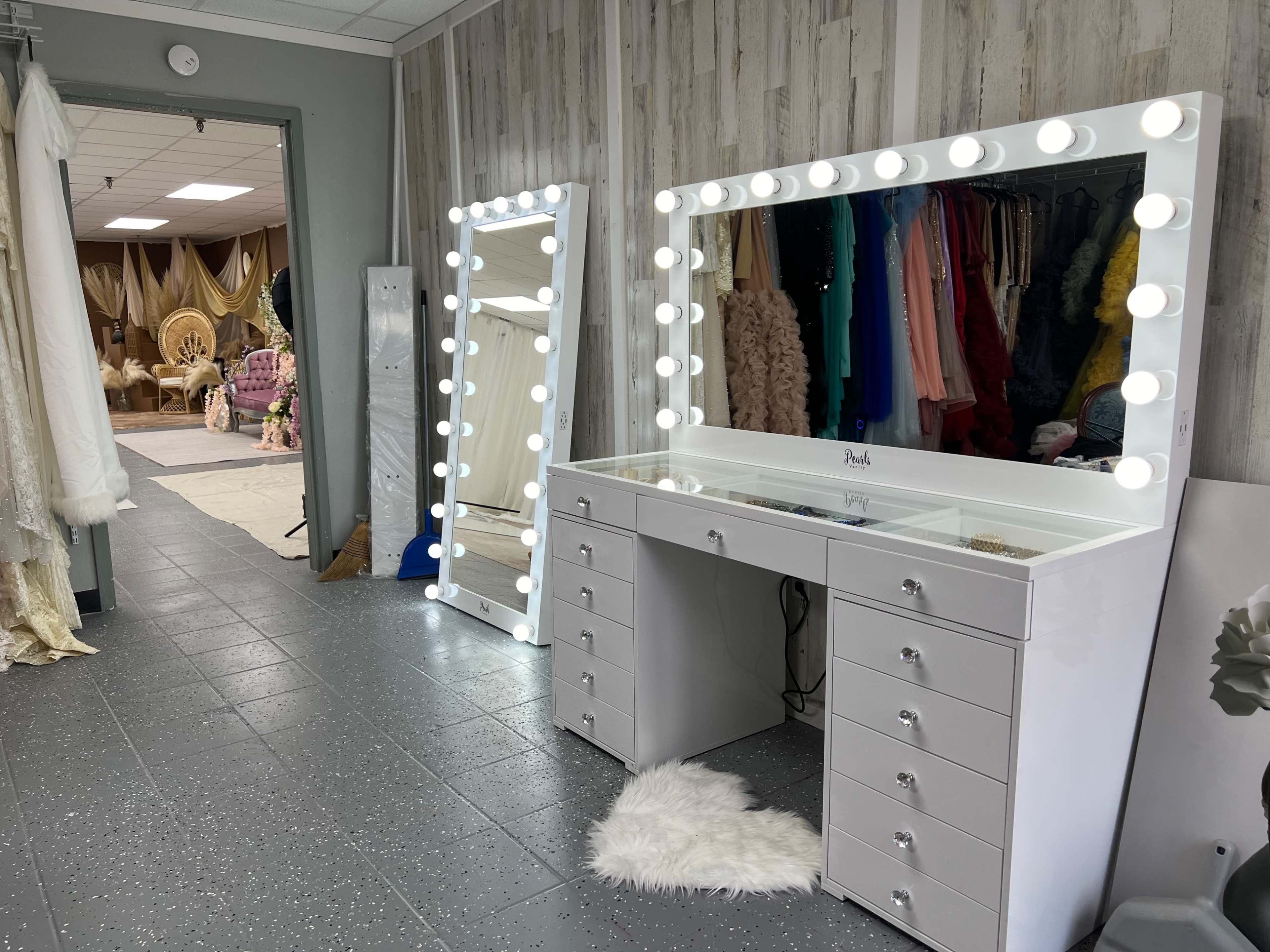 The image shows a well-lit vanity table with a large mirror surrounded by light bulbs, situated in a room that includes clothing racks and a plush area in the background.