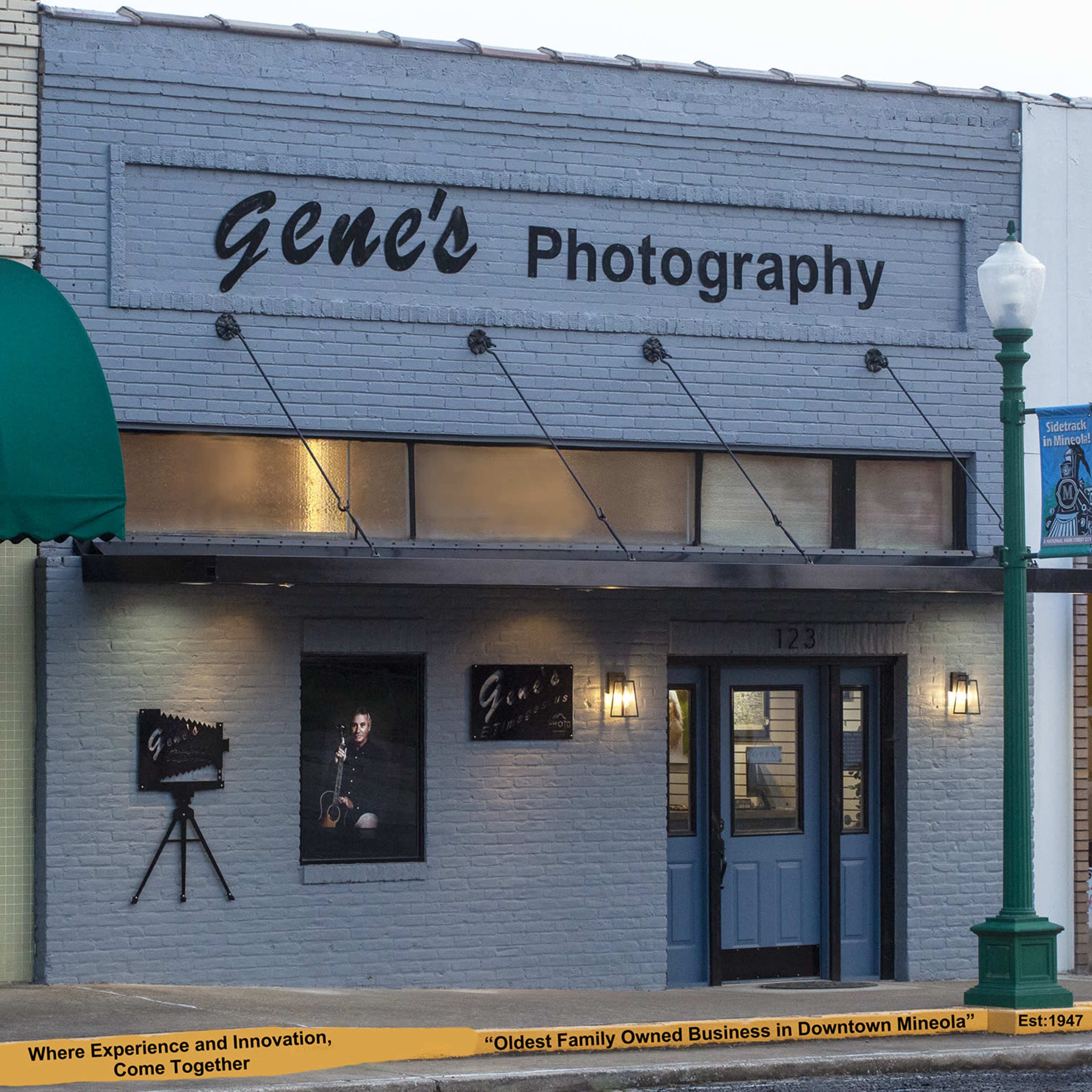The storefront of Gene's Photography features a modern design with large windows showcasing photographs and a sign above that highlights its status as the oldest family-owned business in downtown Mineola, established in 1947.
